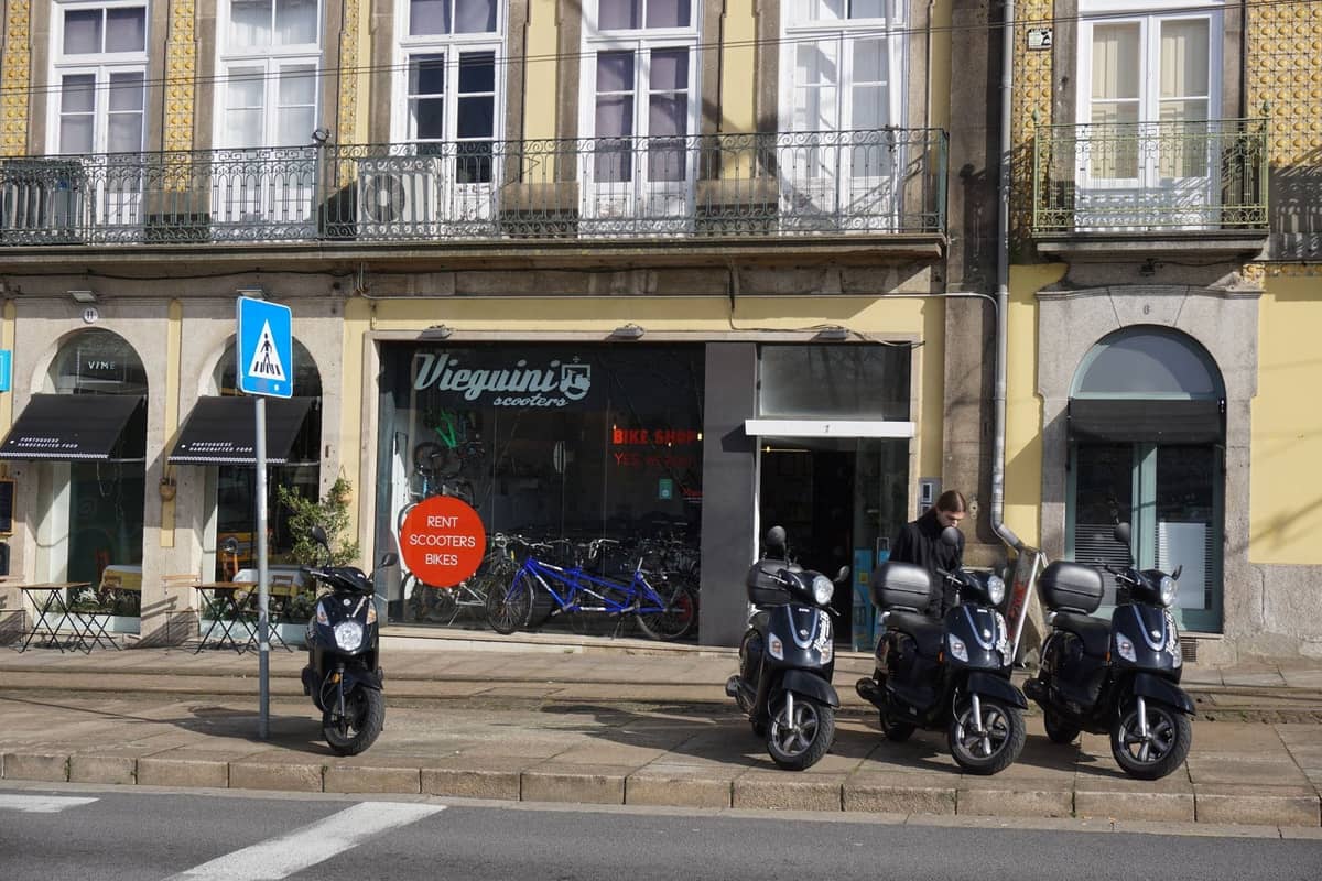 Reihe von Rollern, die vor einem lokalen Geschäft und Café in Porto geparkt sind, mit klassischer gelber portugiesischer Architektur und Balkonen darüber.