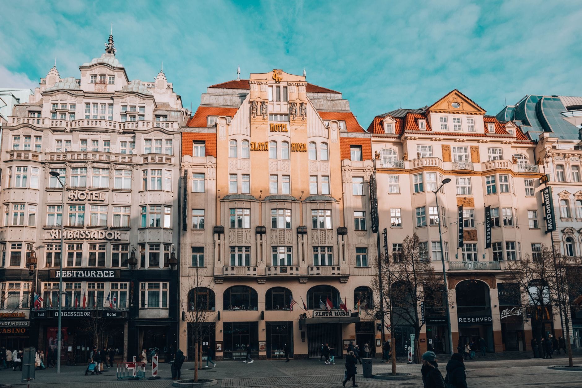 Prächtige historische Gebäude an einer belebten Straße im Zentrum von Prag mit einem Hoteleingang, Geschäften und Passanten unter strahlend blauem Himmel.