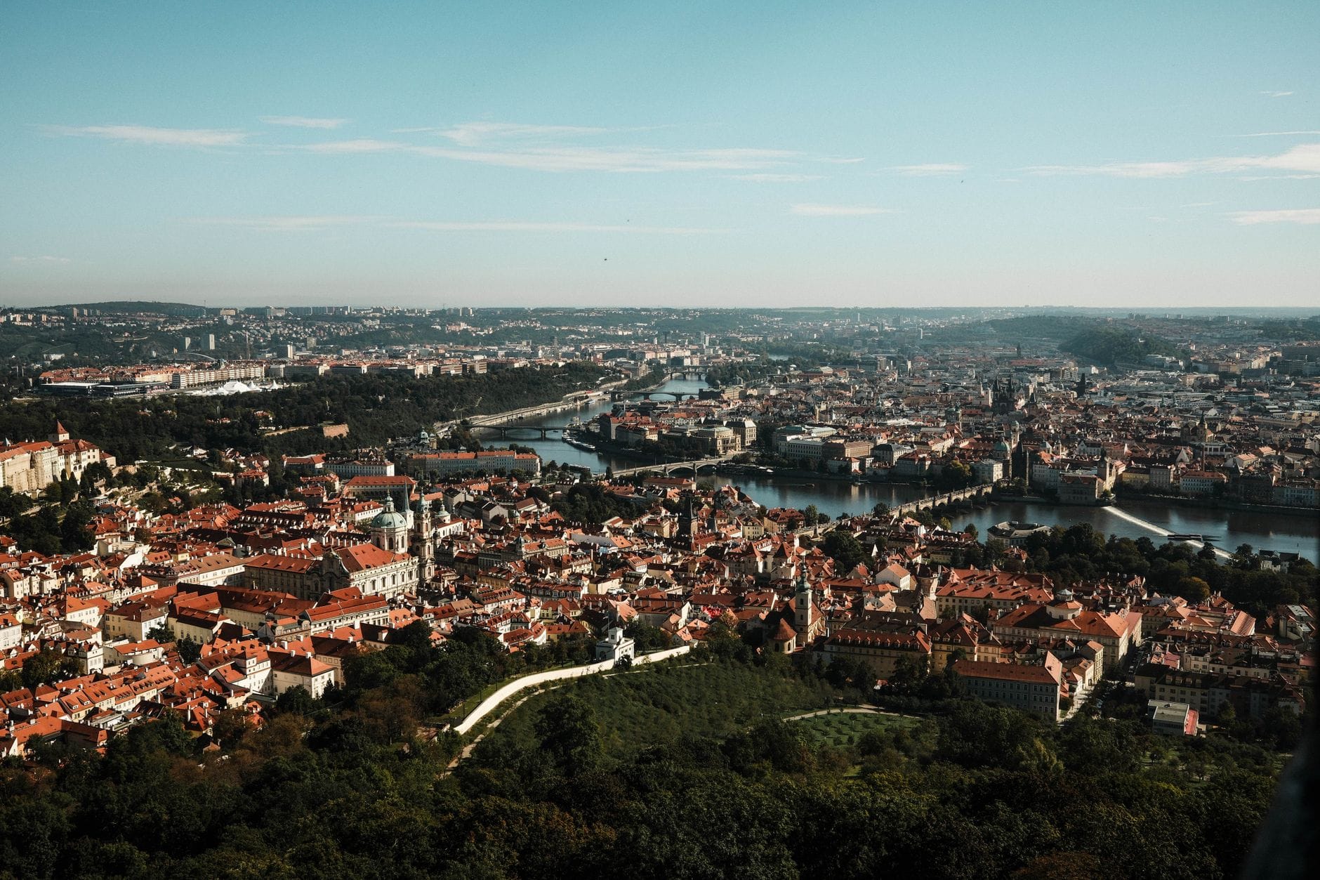 Szeroka panoramiczna fotografia lotnicza Pragi przedstawiająca rzekę Wełtawę, historyczne dzielnice miasta, budynki z czerwonymi dachami i liczne mosty łączące oba brzegi.