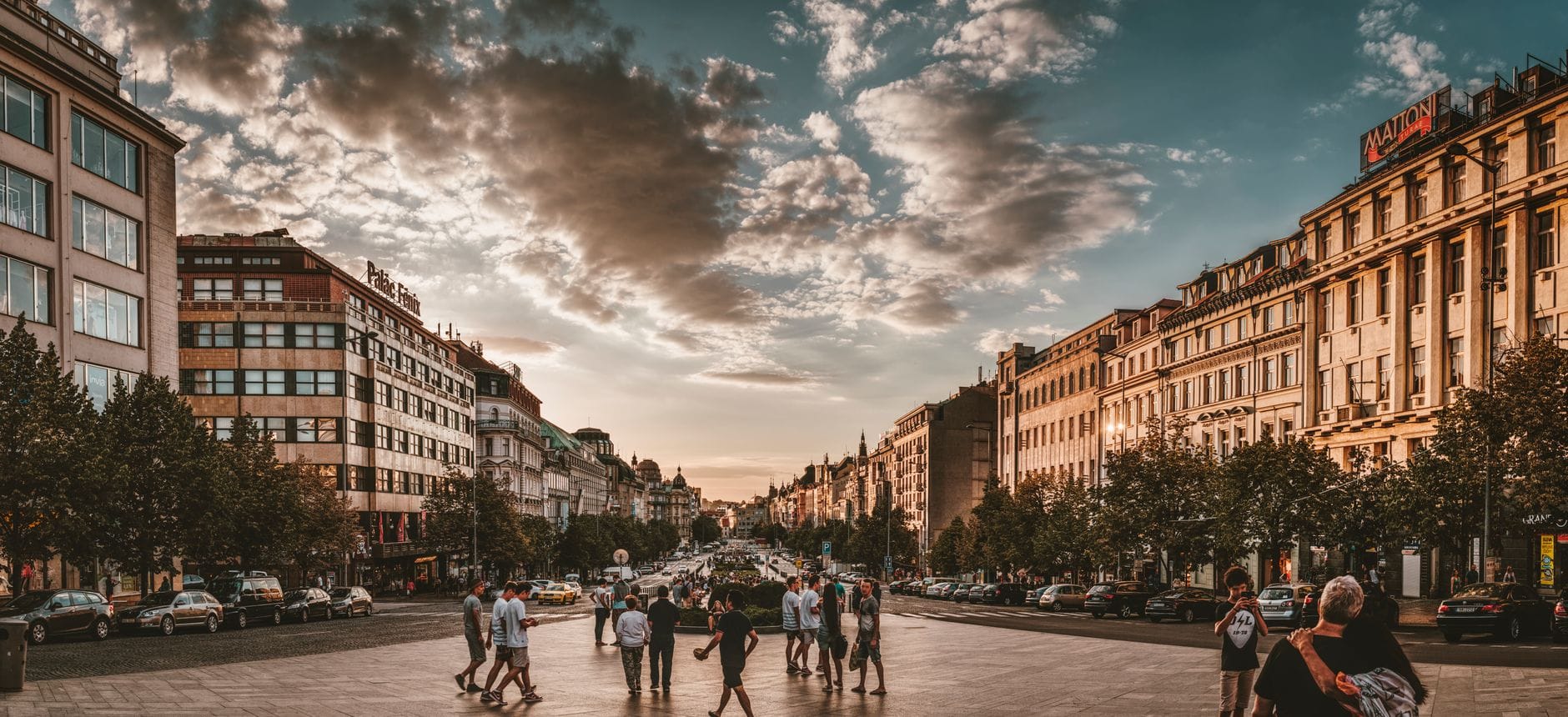 Szeroki widok na Plac Wacława w praskiej Nowej Części Miasta, otoczony historycznymi budynkami i tętniący ruchem pieszych i samochodów pod dramatycznym niebem.