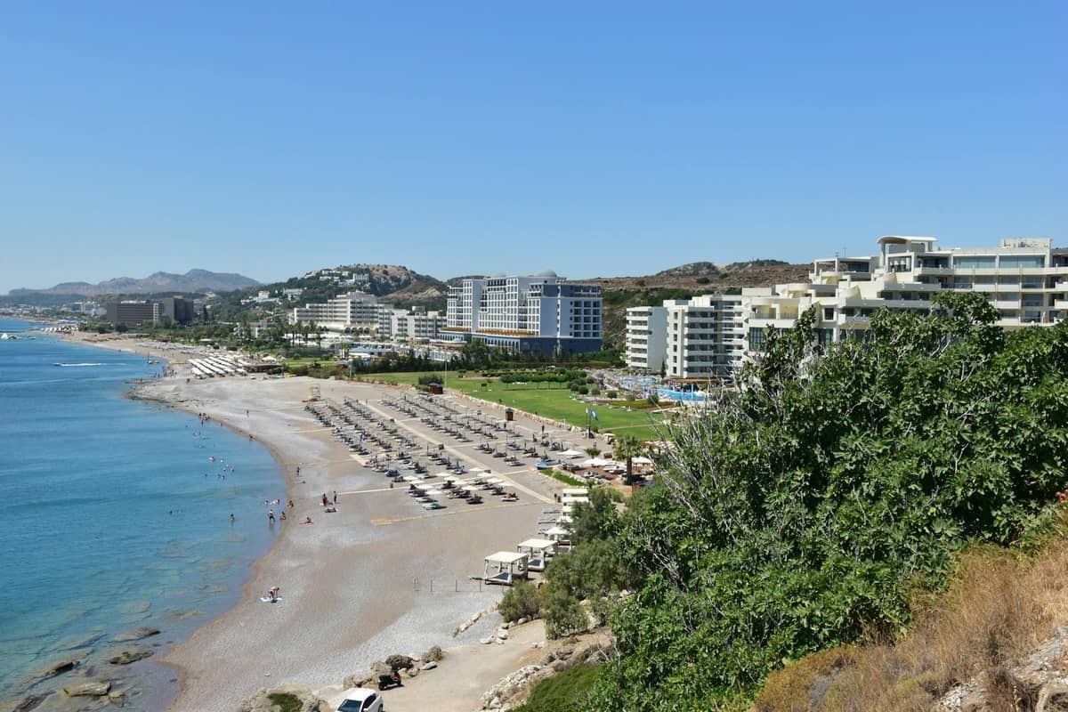 Long sandy beach lined with sun loungers and umbrellas, with large resort hotels in the background along Rhodes' east coast.