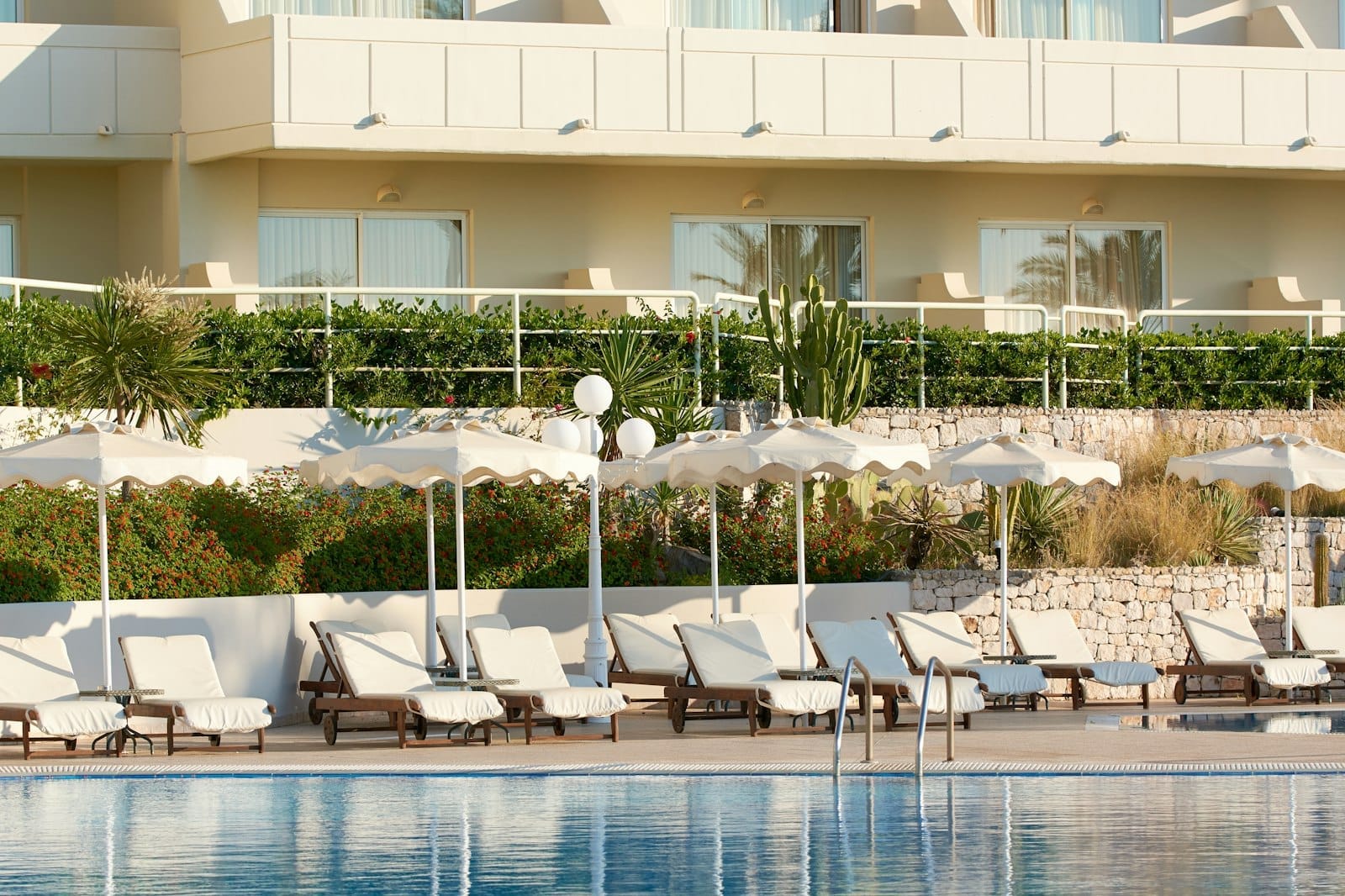 Hotel moderno em Rodes com varandas com vista para uma área de piscina ajardinada, espreguiçadeiras, guarda-sóis brancos e plantas mediterrâneas num dia ensolarado.