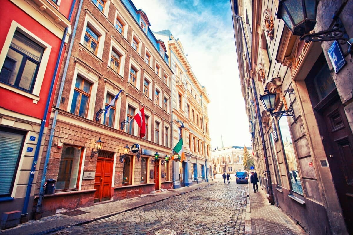 Bunte historische Gebäude mit lettischen und irischen Flaggen säumen eine Kopfsteinpflasterstraße in der Rigaer Altstadt an einem klaren, hellen Tag.