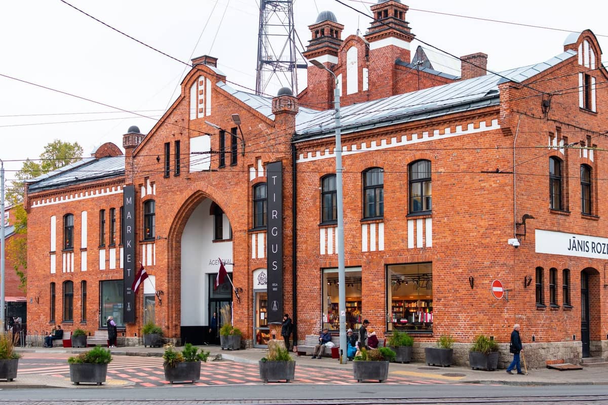 Historisches rotes Backsteingebäude des Āgenskalns-Markts in Riga, mit vorbeigehenden Menschen und lettischen Flaggen an der Fassade.