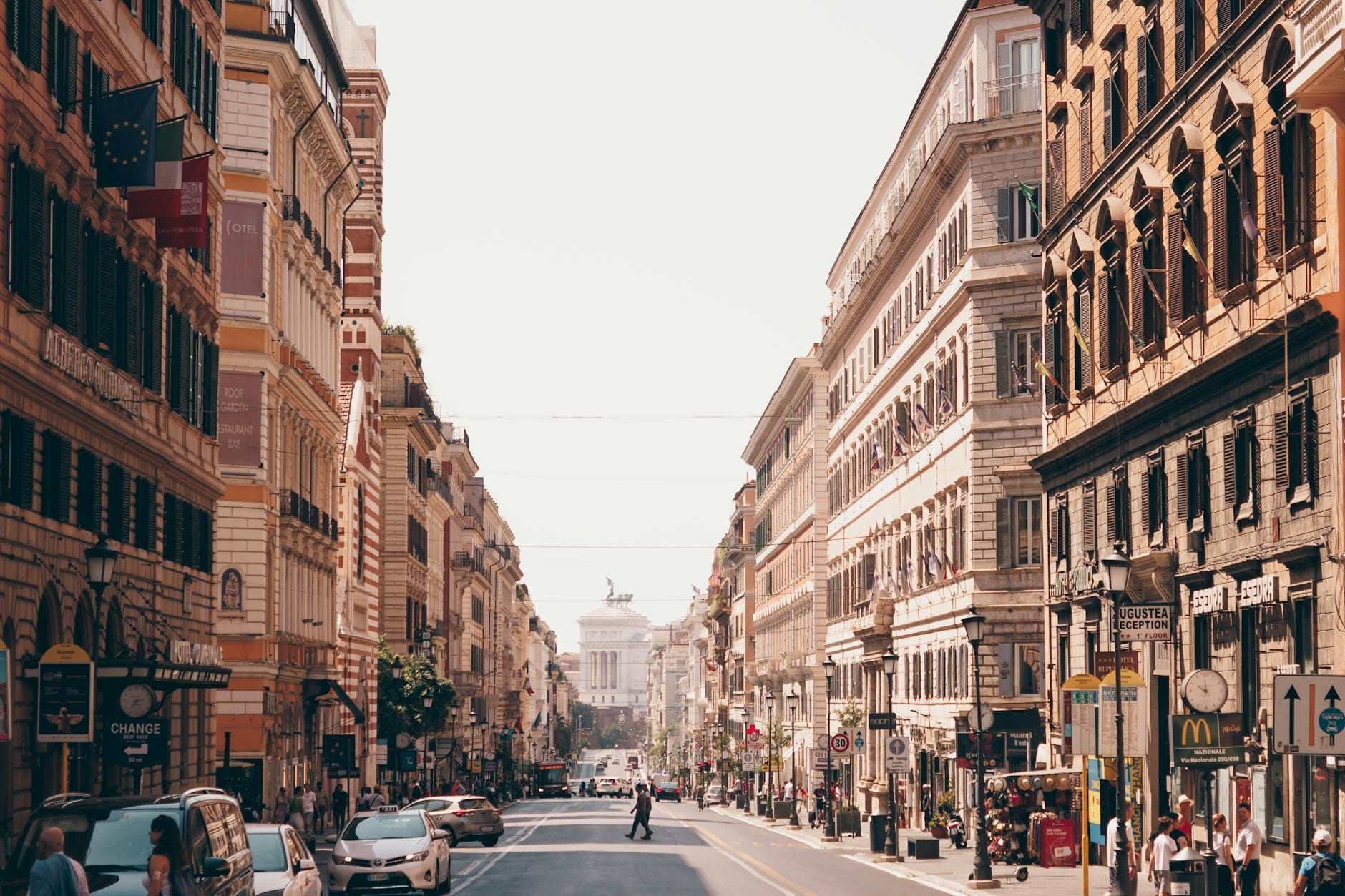 Broad street in the Monti neighborhood of Rome with classic buildings, some traffic, and the Vittoriano monument visible in the distance.