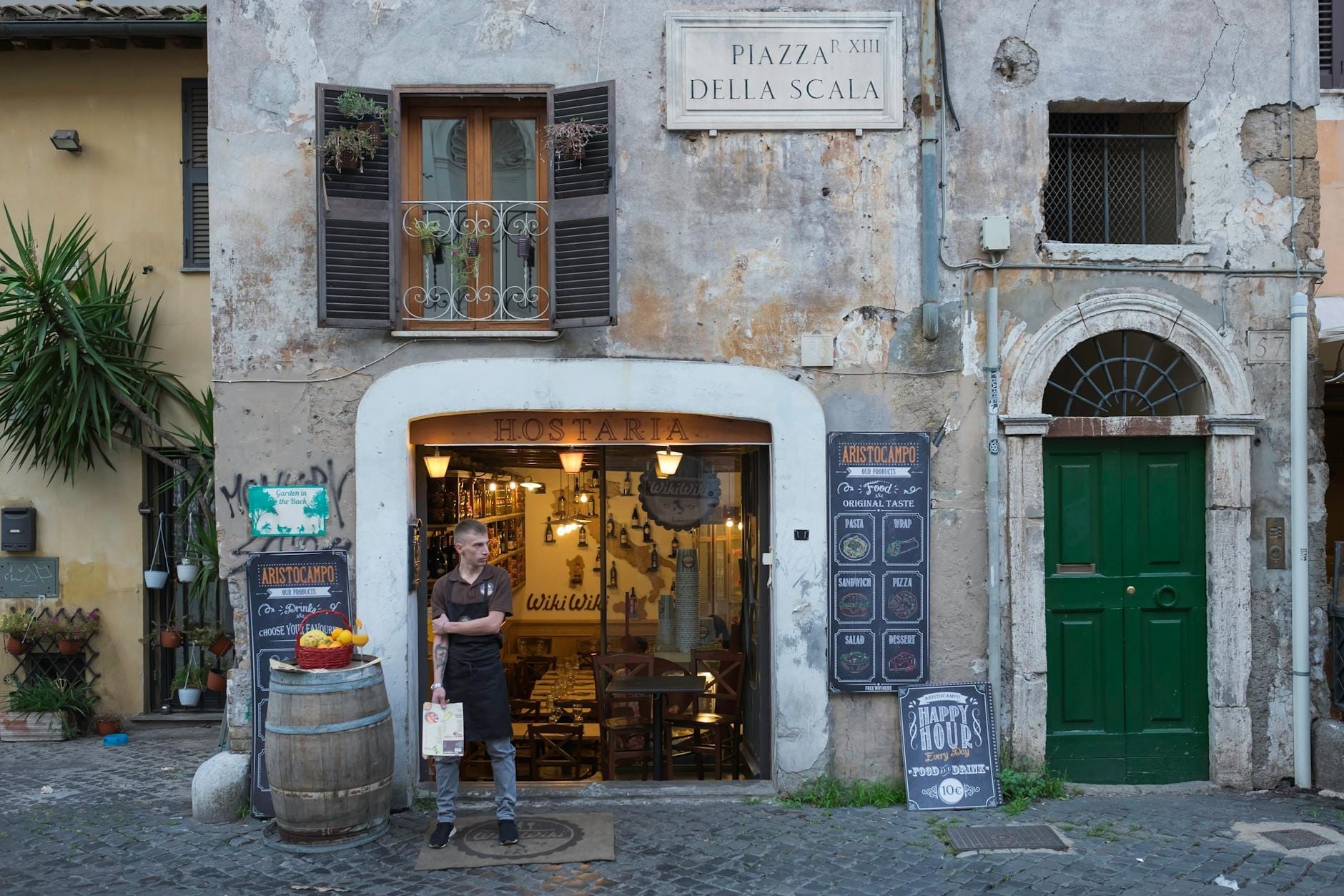 Charming rustic building with Piazza della Scala sign, cozy trattoria entrance, and a person standing outside on a cobblestone street in Trastevere, Rome.