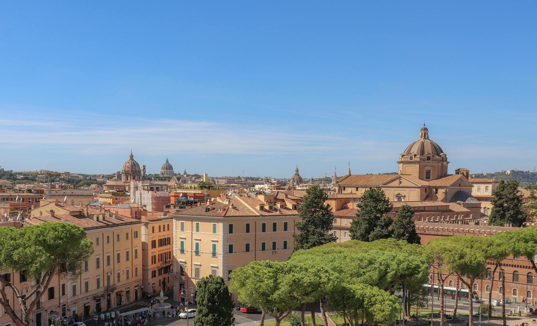 Panoramic view of Rome cityscape with domed churches, classic Roman buildings, and green trees beneath a bright blue sky, showcasing several neighborhoods.