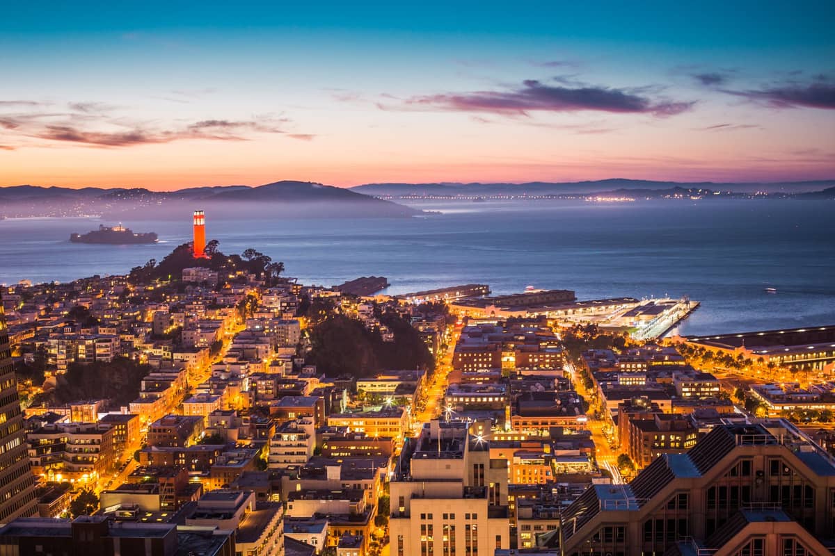 Vista aérea de San Francisco al atardecer con los barrios iluminados, la Torre Coit y la bahía al fondo.
