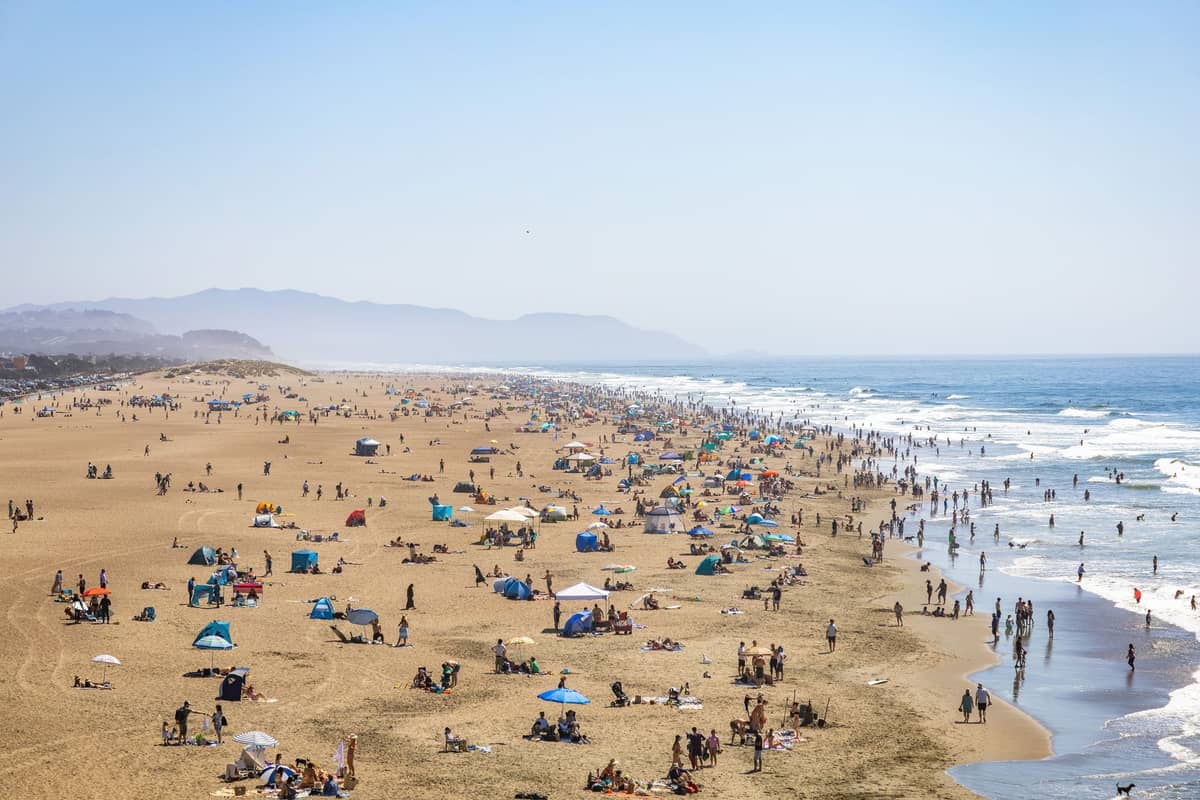 Menschenmassen genießen den Ocean Beach in San Francisco an einem sonnigen Tag, mit Wellen, Sand und Bergen im Hintergrund.