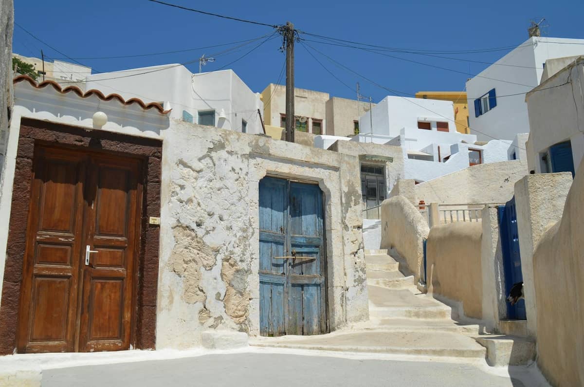 Sunny narrow street in Santorini’s inland village with traditional whitewashed buildings, old wooden doors, and stone steps winding uphill.