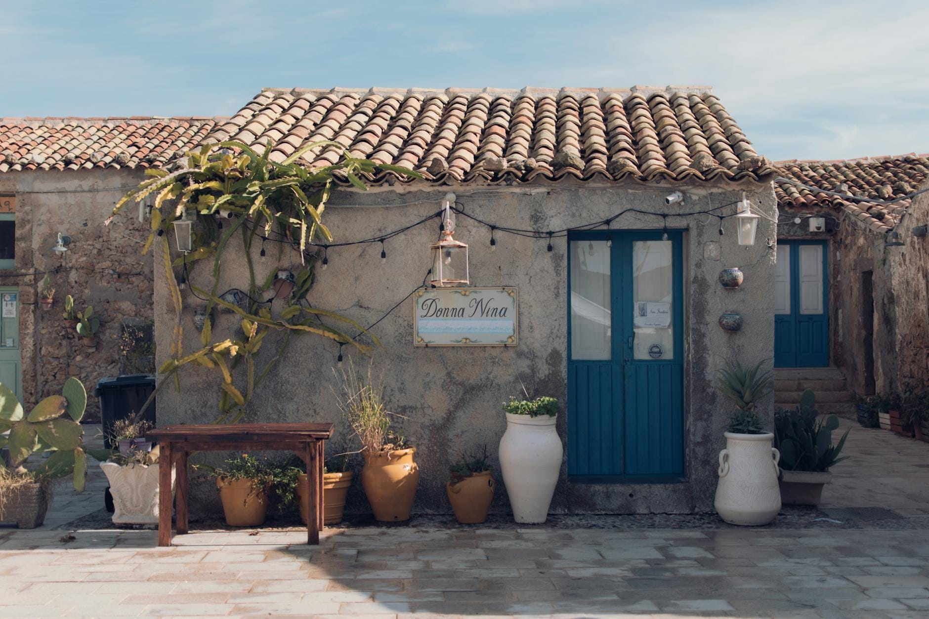 Rustikales Gästehaus auf Sardegna mit Steinmauern, blauen Türen, Topfpflanzen und einem Schild, das den lokalen Unterkunftsstil repräsentiert.