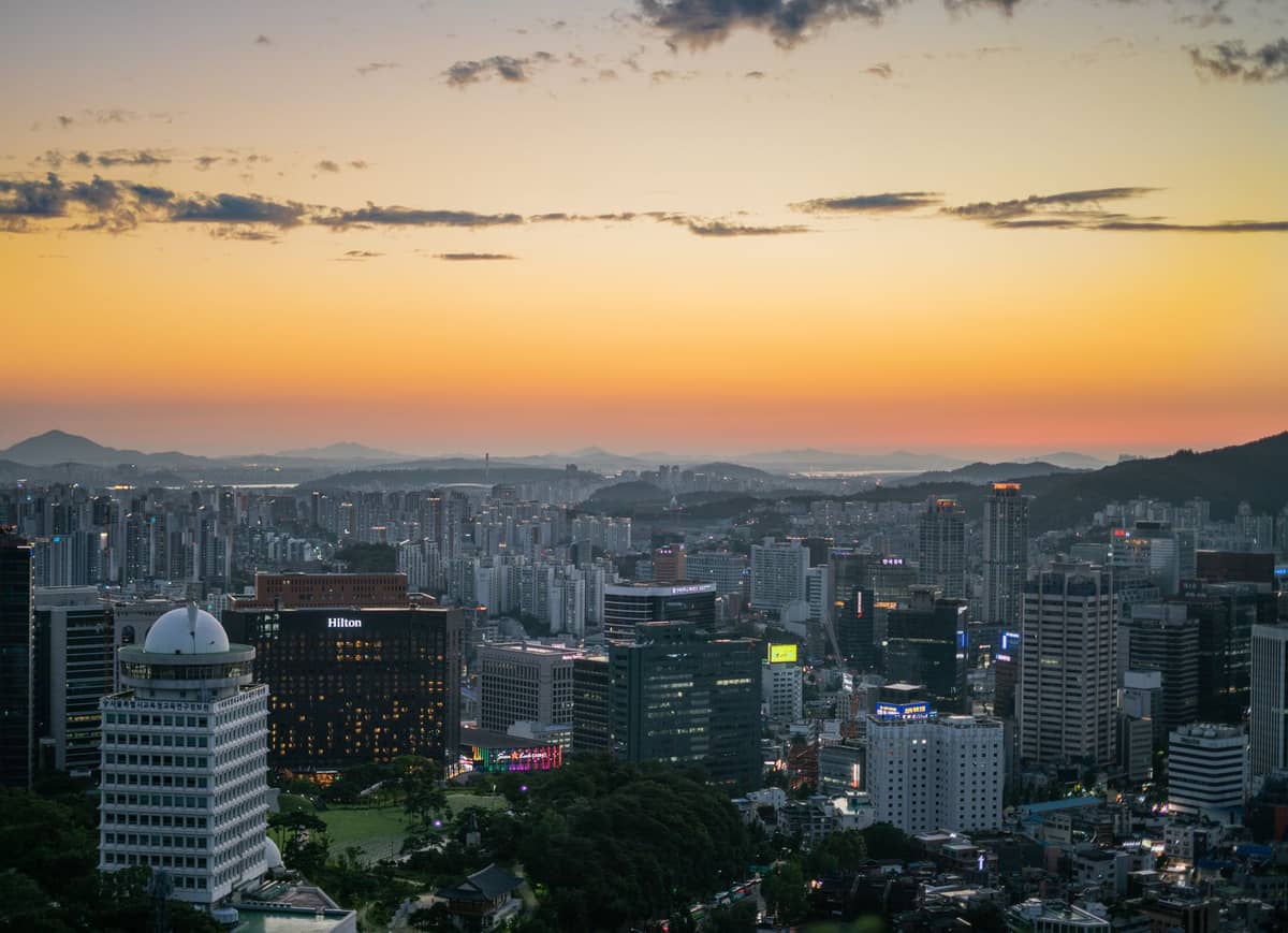 Vista panorâmica do horizonte de Seul ao pôr do sol, com muitos prédios, luzes da cidade e colinas ao fundo sob um céu colorido.
