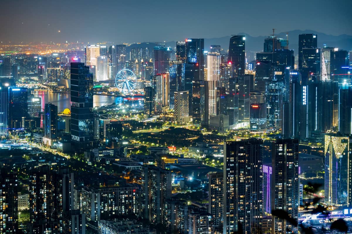 Um vibrante panorama noturno do skyline moderno de Shenzhen, com arranha-céus imponentes, iluminação dramática e o calçadão à beira-mar iluminado, mostrando claramente a paisagem urbana da cidade.