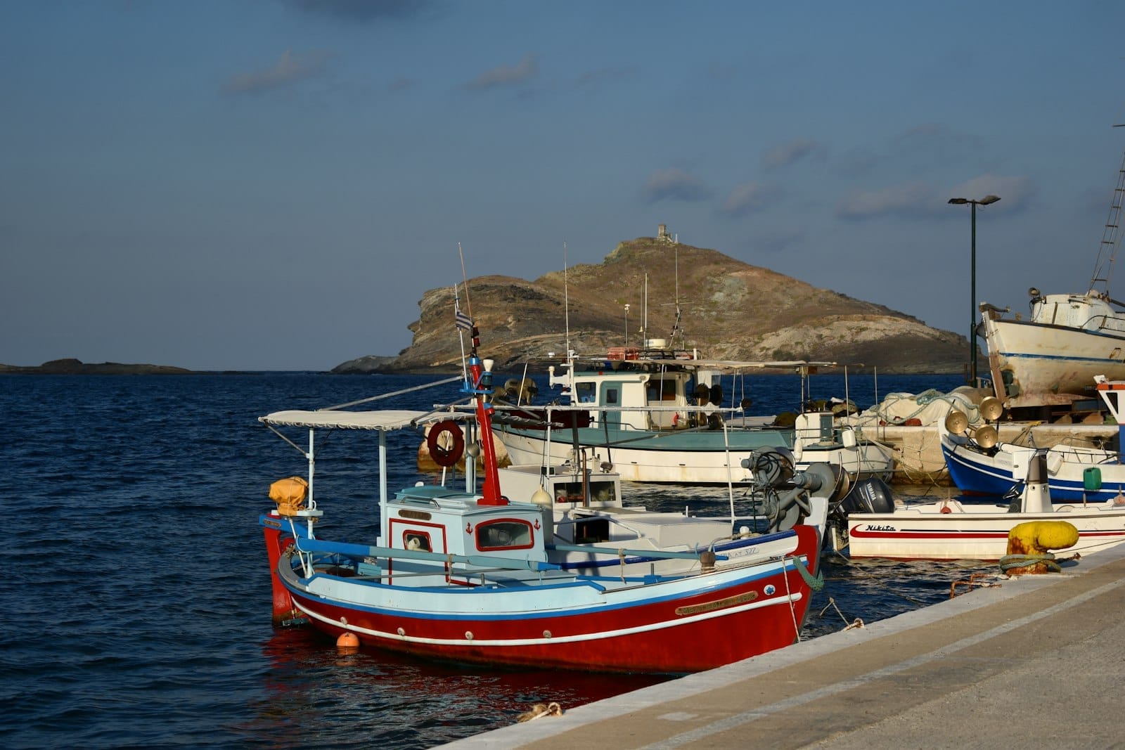 Bunte Fischerboote in einem kleinen Hafen mit einer felsigen Insel im Hintergrund und ruhigem Meer unter blauem Himmel.