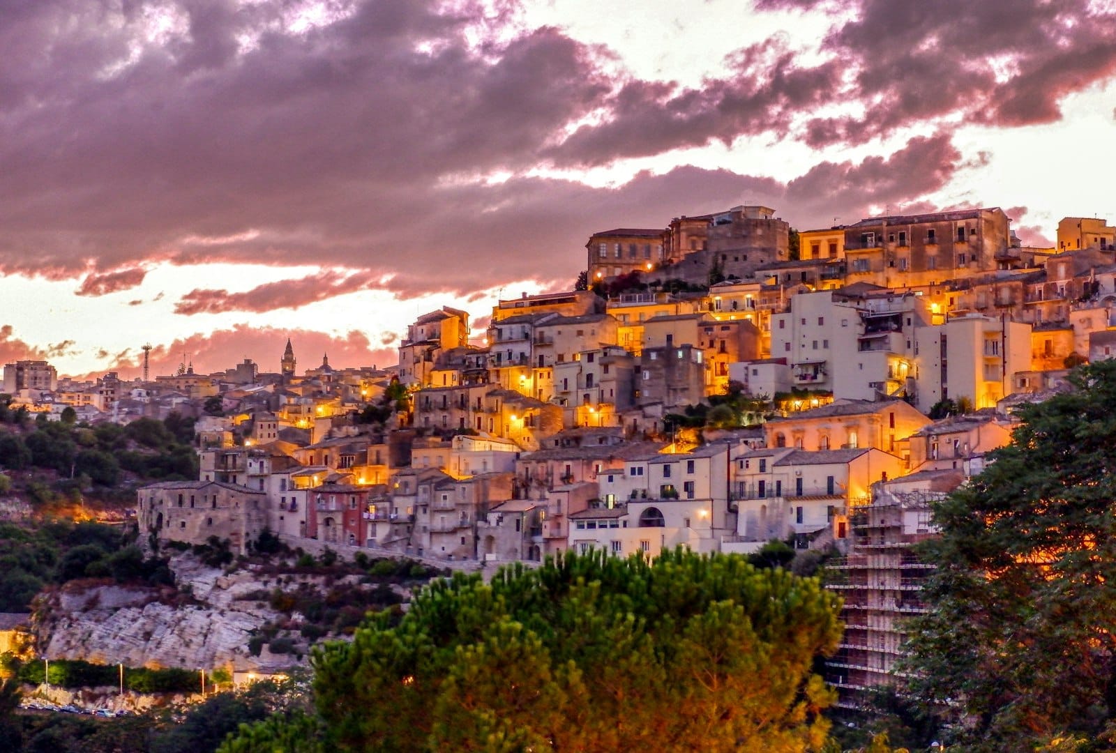 Bunte Hanggebäude von Ragusa auf Sizilien in der Dämmerung beleuchtet, mit dramatischen Wolken und warmem Licht, das eine malerische mediterrane Szene schafft.