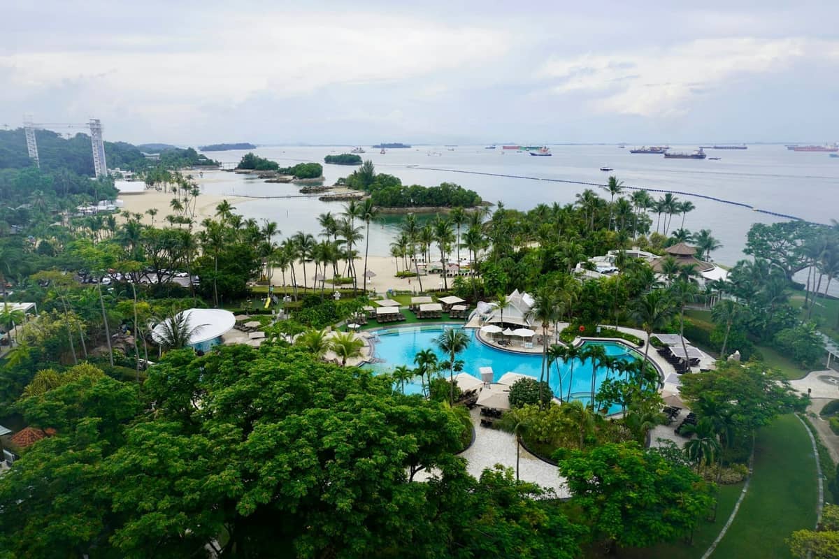Aerial view of Sentosa beachfront with lush greenery, a resort pool, sandy beaches, and ships anchored offshore under a cloudy sky.