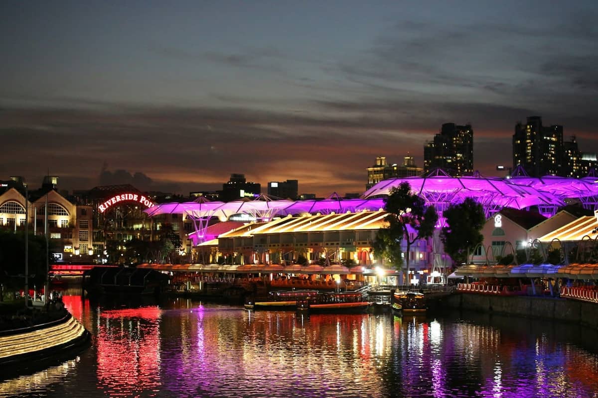 Vibrant night scene of Clarke Quay Singapore, with colorful lights reflecting on the river, bustling riverfront restaurants, and city buildings in the background.