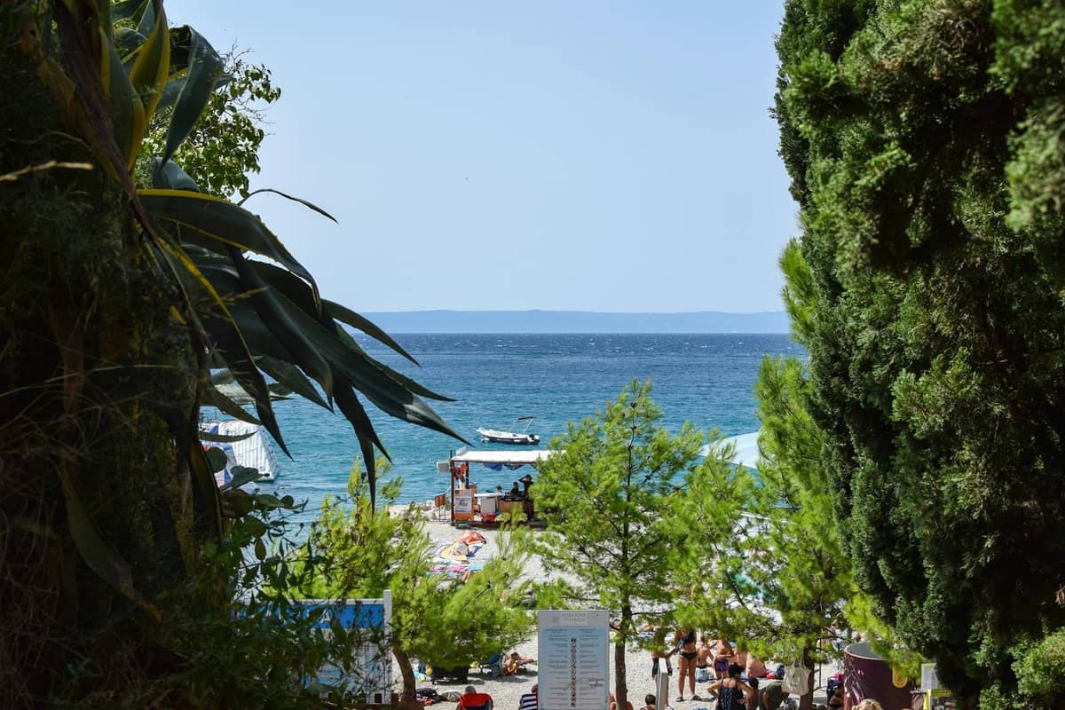 Blick durch Bäume auf einen belebten, mit Sonnenbadenden gefüllten Strand und Badebereich mit blauem Meer und einem kleinen Boot in der Ferne.