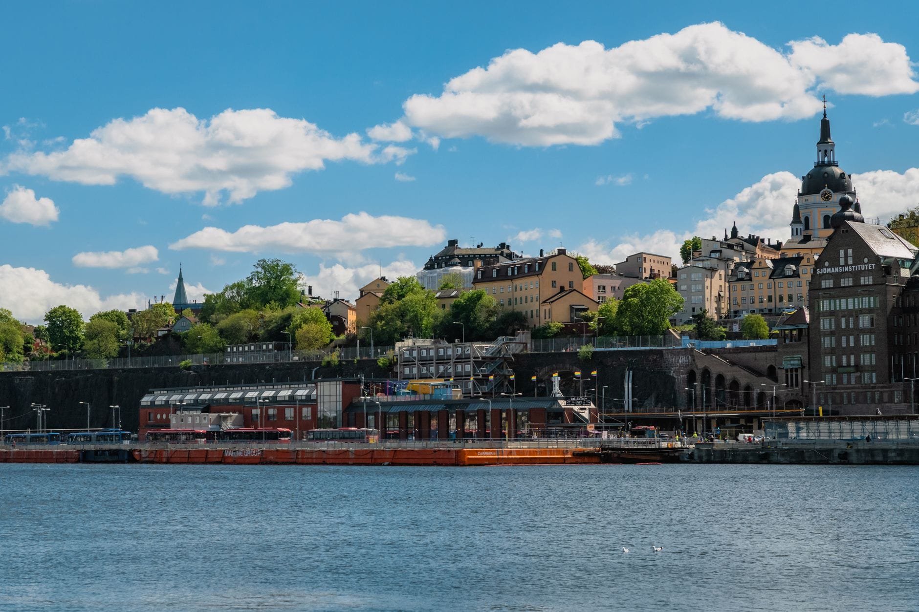 ภาพถ่ายทิวทัศน์ริมน้ำ Södermalm พร้อมอาคารประวัติศาสตร์ ต้นไม้ร่มรื่น และยอดแหลมโบสถ์เหนือผืนน้ำใต้ท้องฟ้าสีฟ้า