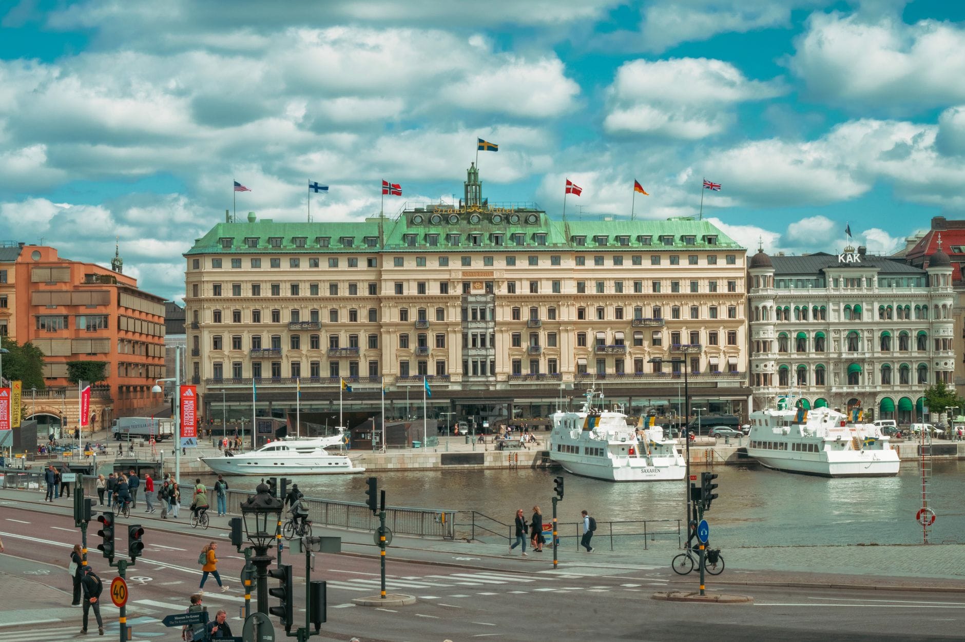 วิวกว้างของ Grand Hôtel Stockholm ริมน้ำ มีเรือจอดและผู้คนเดินเล่นริมท่าเรือด้านหน้าโรงแรม