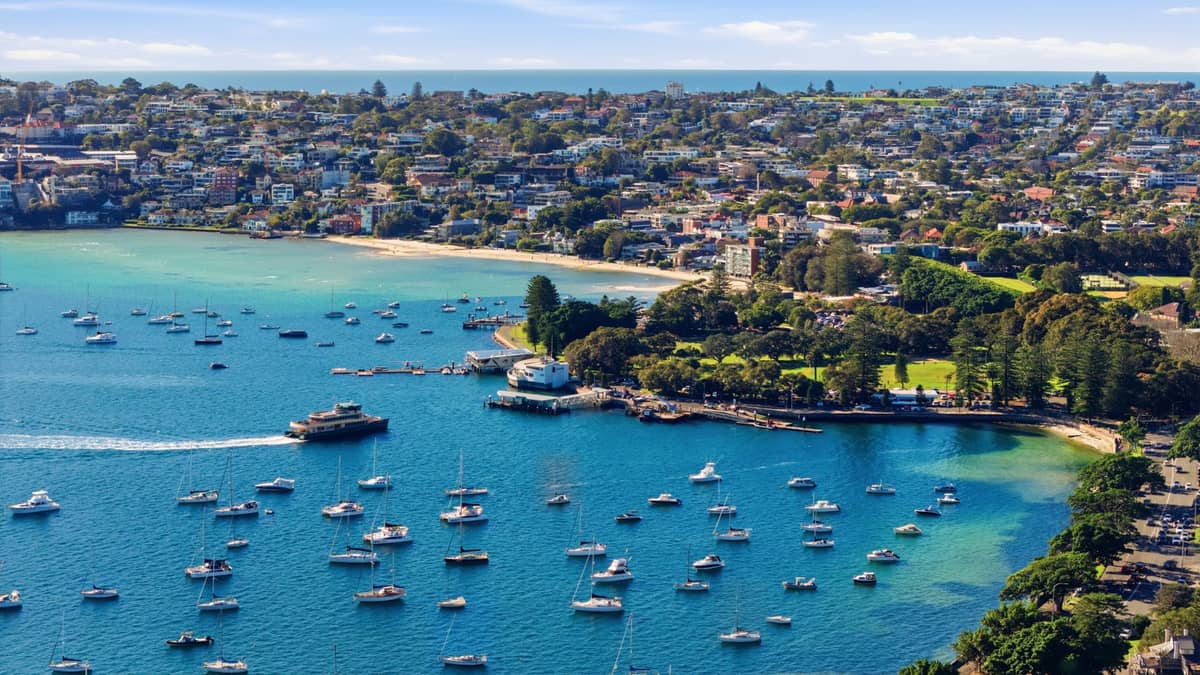 Aerial view of Manly showing boats in the harbor, the ferry wharf, sandy beachfront, and residential neighbourhoods on a sunny day.