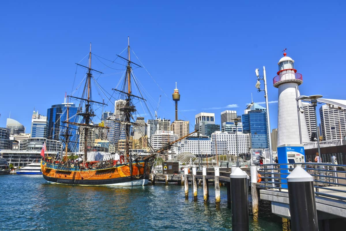 Tall ship docked at Darling Harbour in Sydney with lighthouse, city skyline, and clear blue sky visible in background.