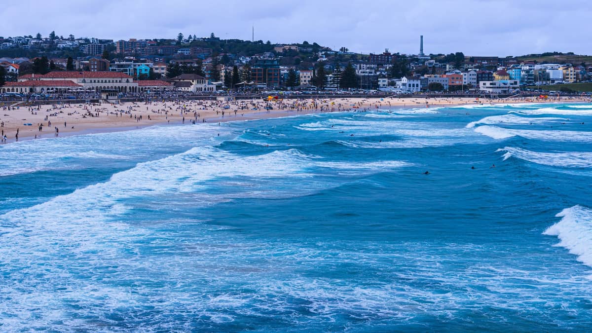 Wide view of Bondi Beach with crowds, blue waves, and the suburb’s distinctive buildings in the background under a cloudy sky.