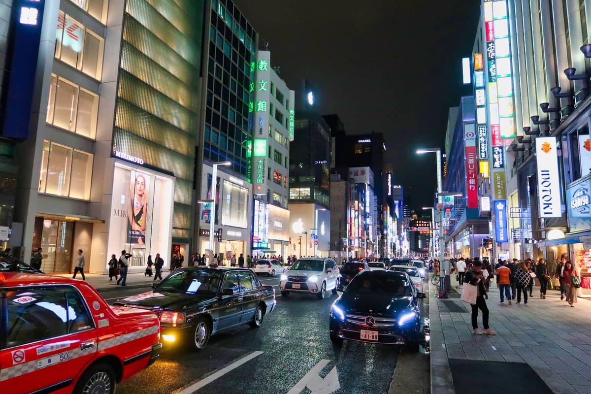 Night view of Ginza's upscale shopping street in Tokyo, lined with luxury boutiques, bright signage, and taxis with people walking on the sidewalk.