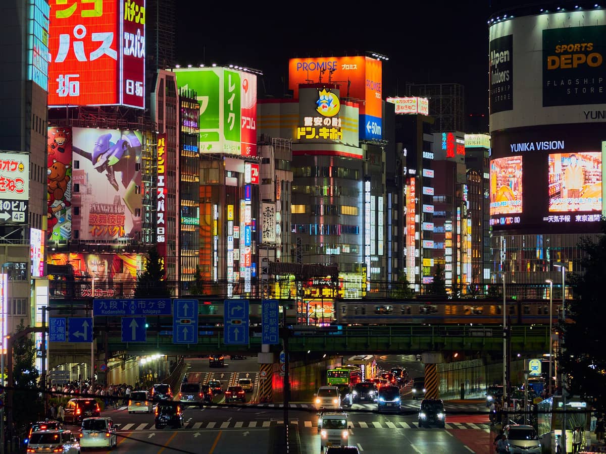 Night view of Shinjuku with bright neon signs, billboards, a busy intersection, and a train passing on an overhead track.