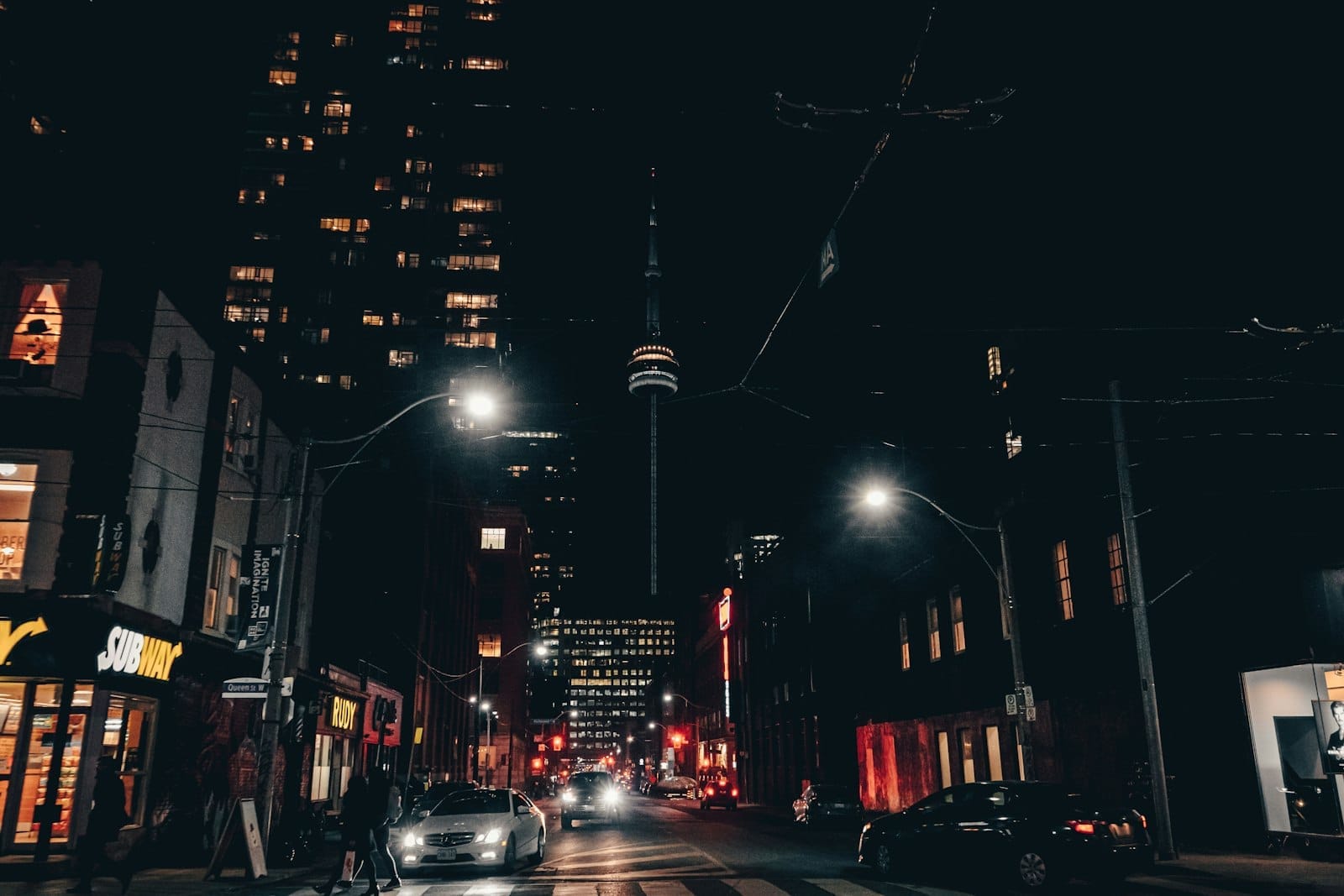 Vista noturna de uma rua no centro de Toronto com a CN Tower visível, cercada por bares, restaurantes e edifícios iluminados.