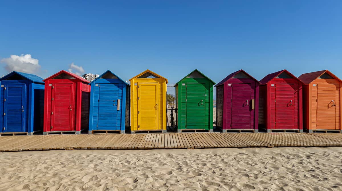 Rangée de cabines de plage colorées sur le sable avec un ciel bleu, évoquant l'atmosphère méditerranéenne typique des quartiers côtiers de Valencia.