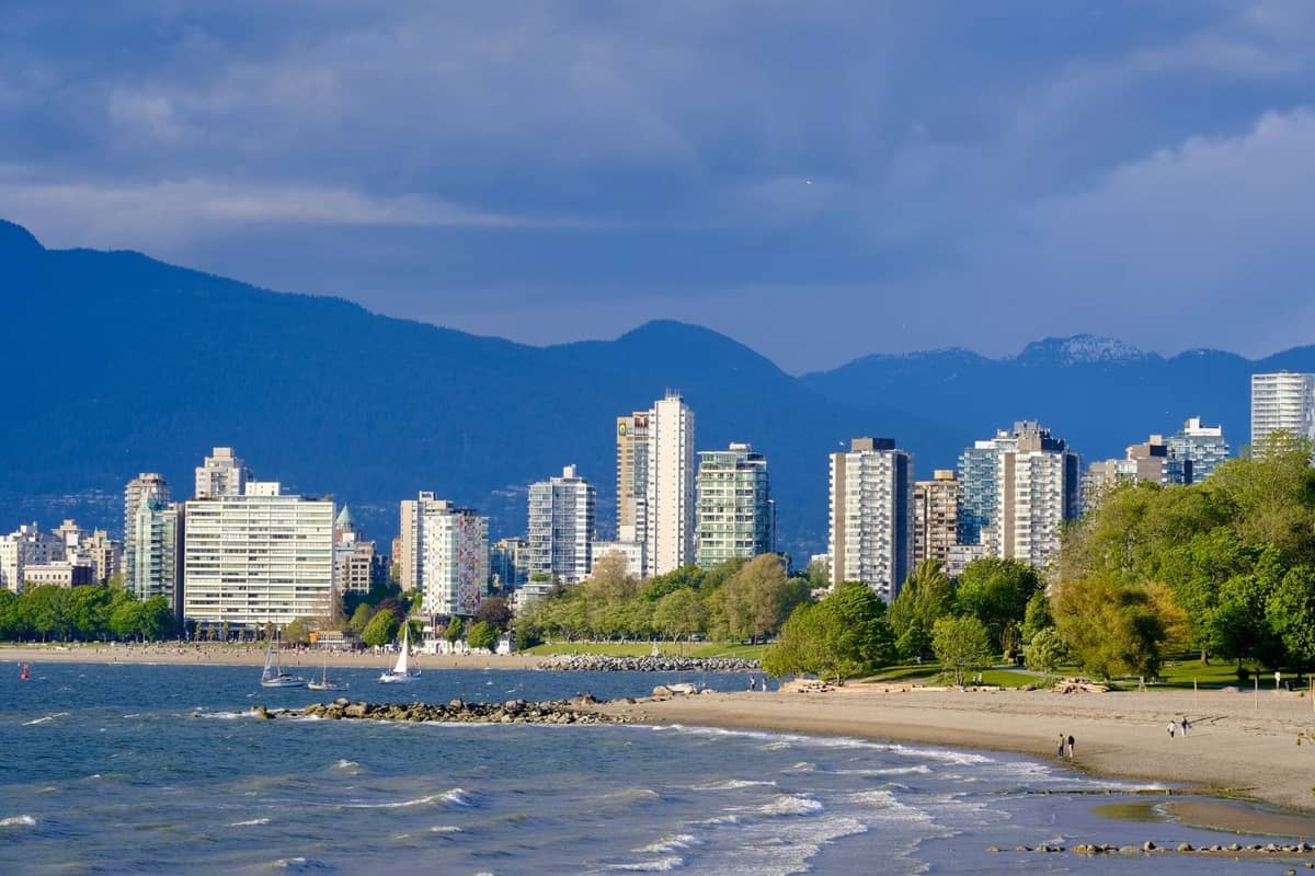 Strand in Vancouver mit Stadtsilhouette, Bergen im Hintergrund und Menschen, die am Wasser über den Sand spazieren.
