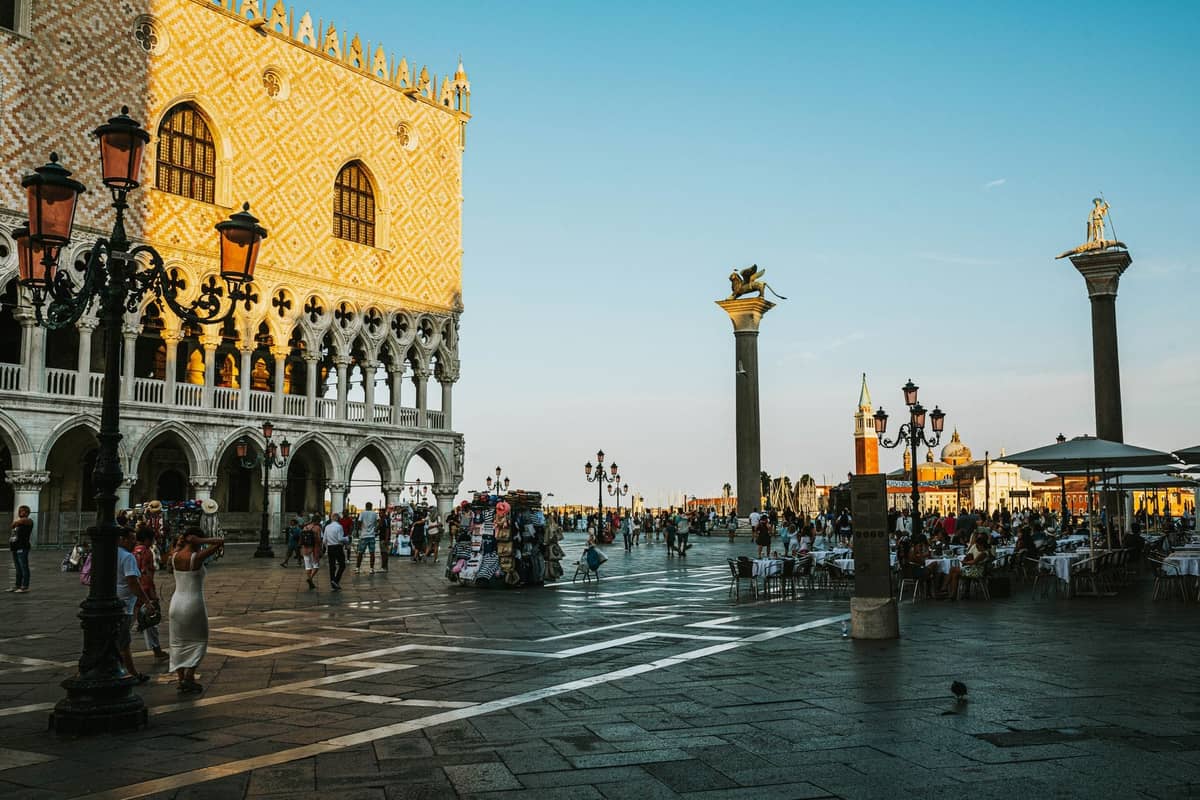 Menschen, die bei Sonnenuntergang auf der belebten Piazzetta San Marco nahe dem Palazzo Ducale spazieren und sitzen, mit Säulen und venezianischer Architektur.