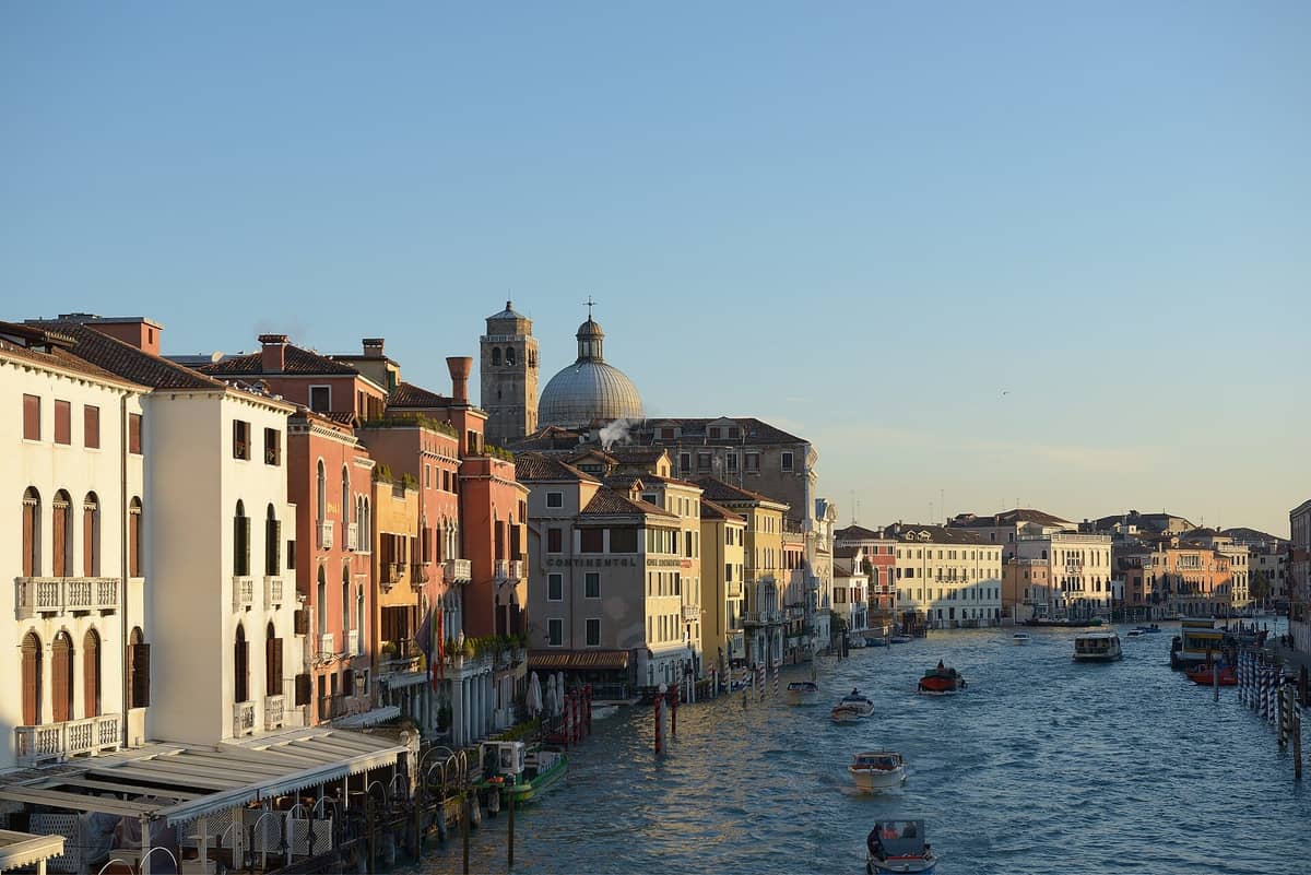 Weitläufiger Blick auf den Canal Grande in Venedig mit bunten historischen Gebäuden entlang der Uferpromenade und Booten, die unter einem klaren Himmel auf dem Wasser fahren.
