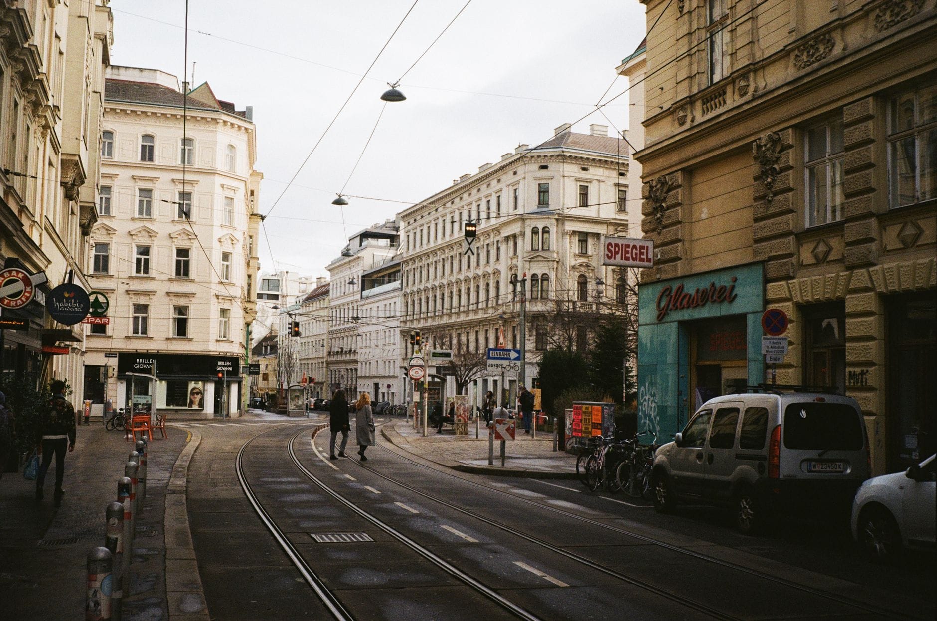 Straßenszene in Wien mit Straßenbahnschienen, historischen Gebäuden, lokalen Geschäften und Fußgängern an einem ruhigen Tag.