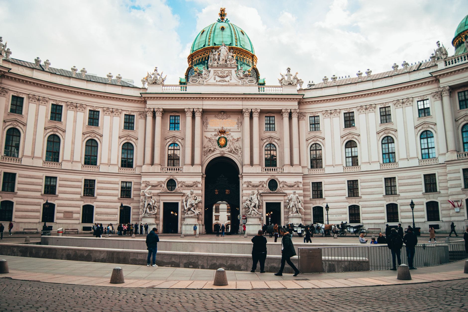 Weitwinkelansicht des Eingangs des Hofburg-Palastes im Innere Stadt in Wien, mit Passanten und aufwendiger Barockarchitektur unter einer grünen Kupferkuppel.