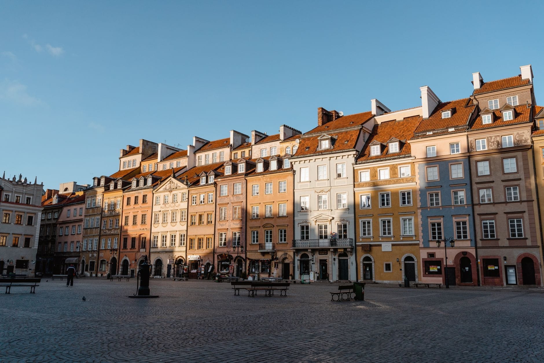 Vista ampla das coloridas casas históricas ao redor de uma praça espaçosa e ensolarada na Cidade Velha de Varsóvia, capturando a atmosfera vibrante e a arquitetura convidativa.