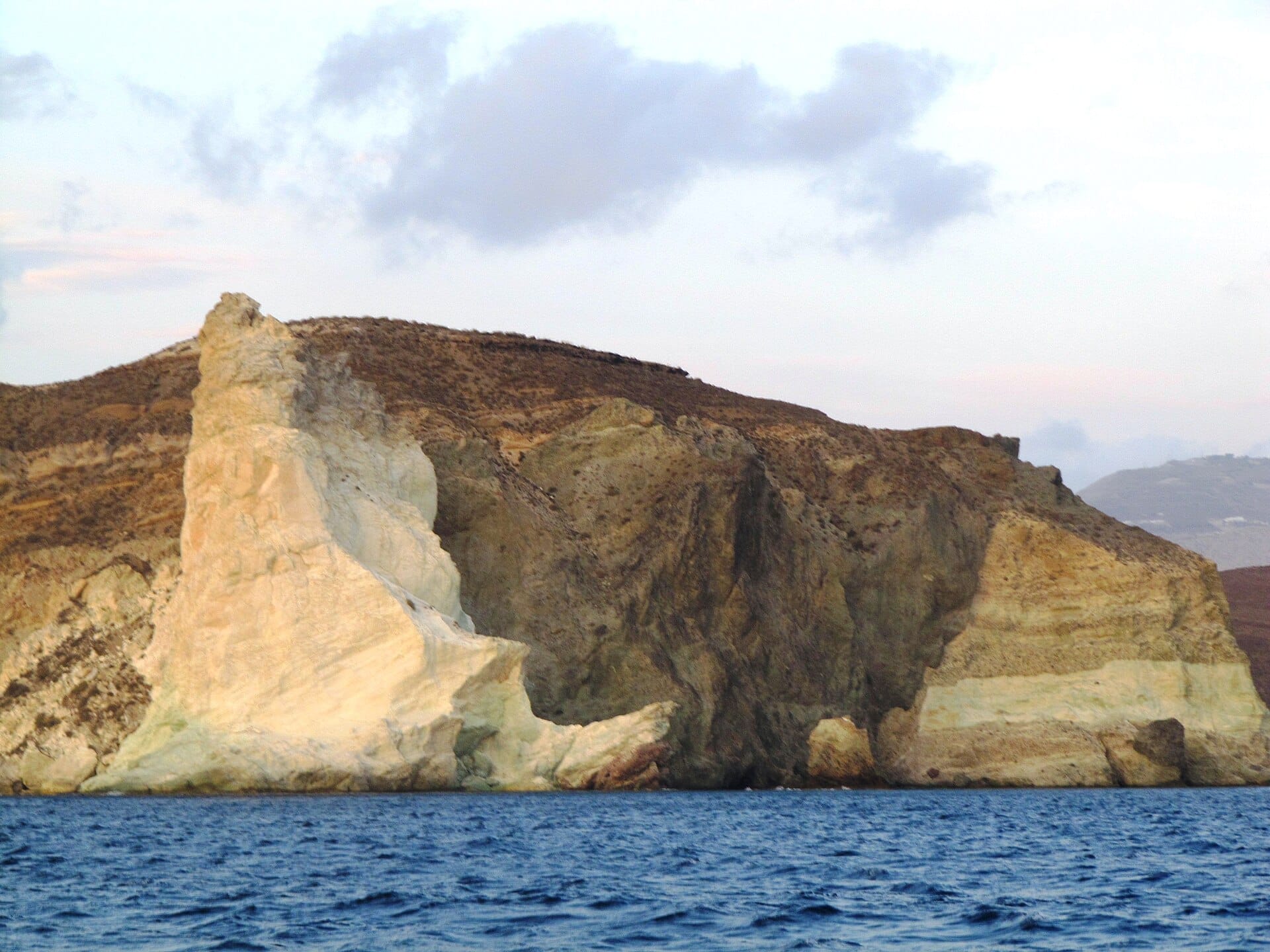 White Beach ที่ Akrotiri, Santorini พร้อมหน้าผาภูเขาไฟสีขาวสูงตระหง่านเหนือทะเล Aegean สีน้ำเงินเข้มใต้ท้องฟ้าที่มีเมฆบางส่วน