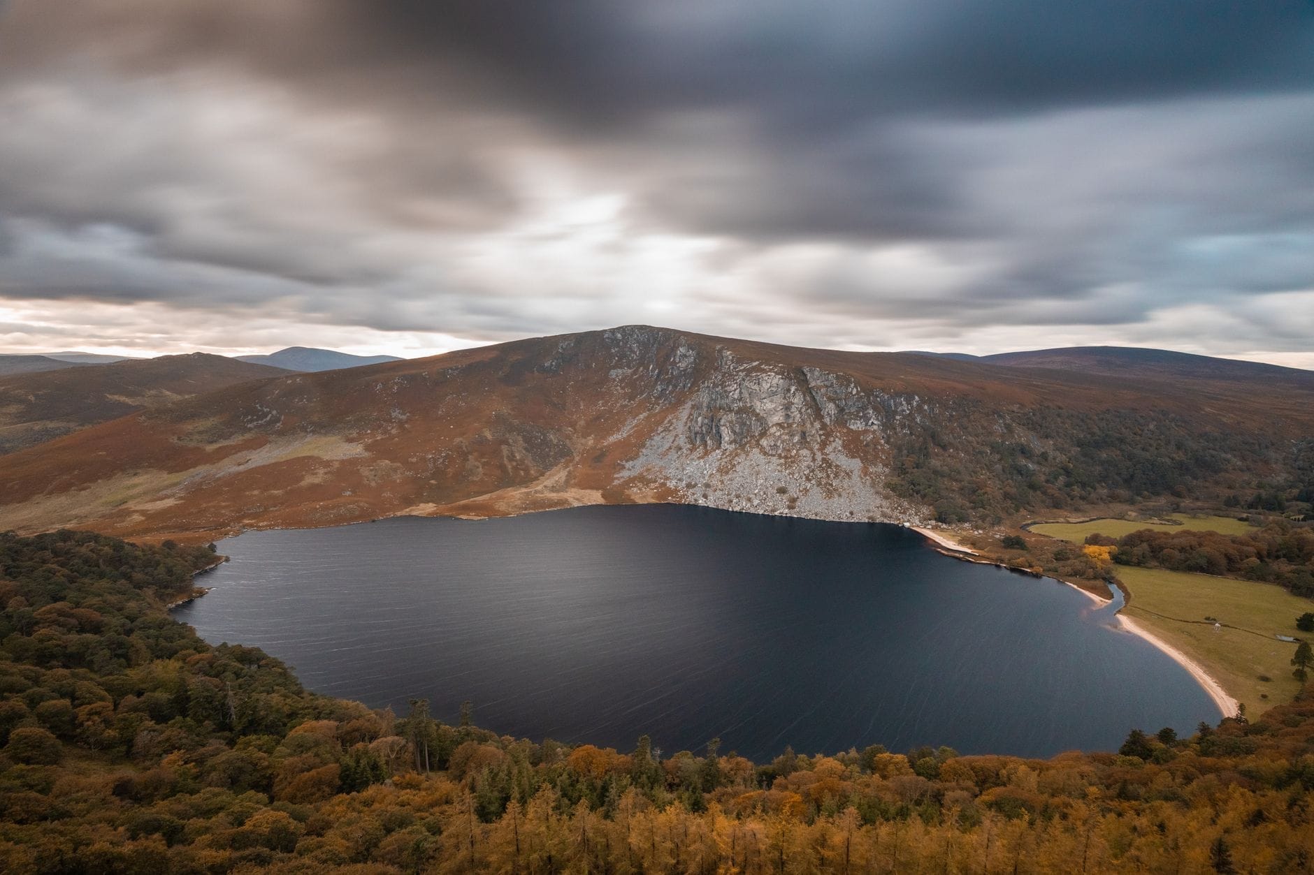 Vista grandangolare spettacolare di Lough Tay con acque scure, rive boscose e colline frastagliate nel Parco Nazionale delle Wicklow Mountains sotto cieli nuvolosi.