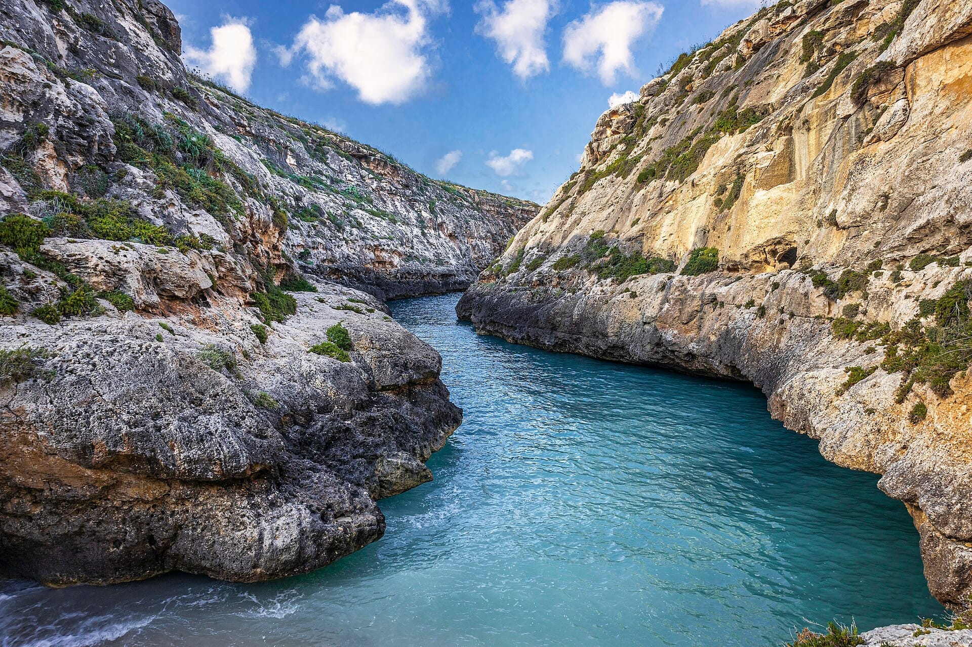 La gola di Wied il-Għasri con acqua turchese che scorre tra scogliere calcaree frastagliate sotto un cielo luminoso sulla costa nord di Gozo.