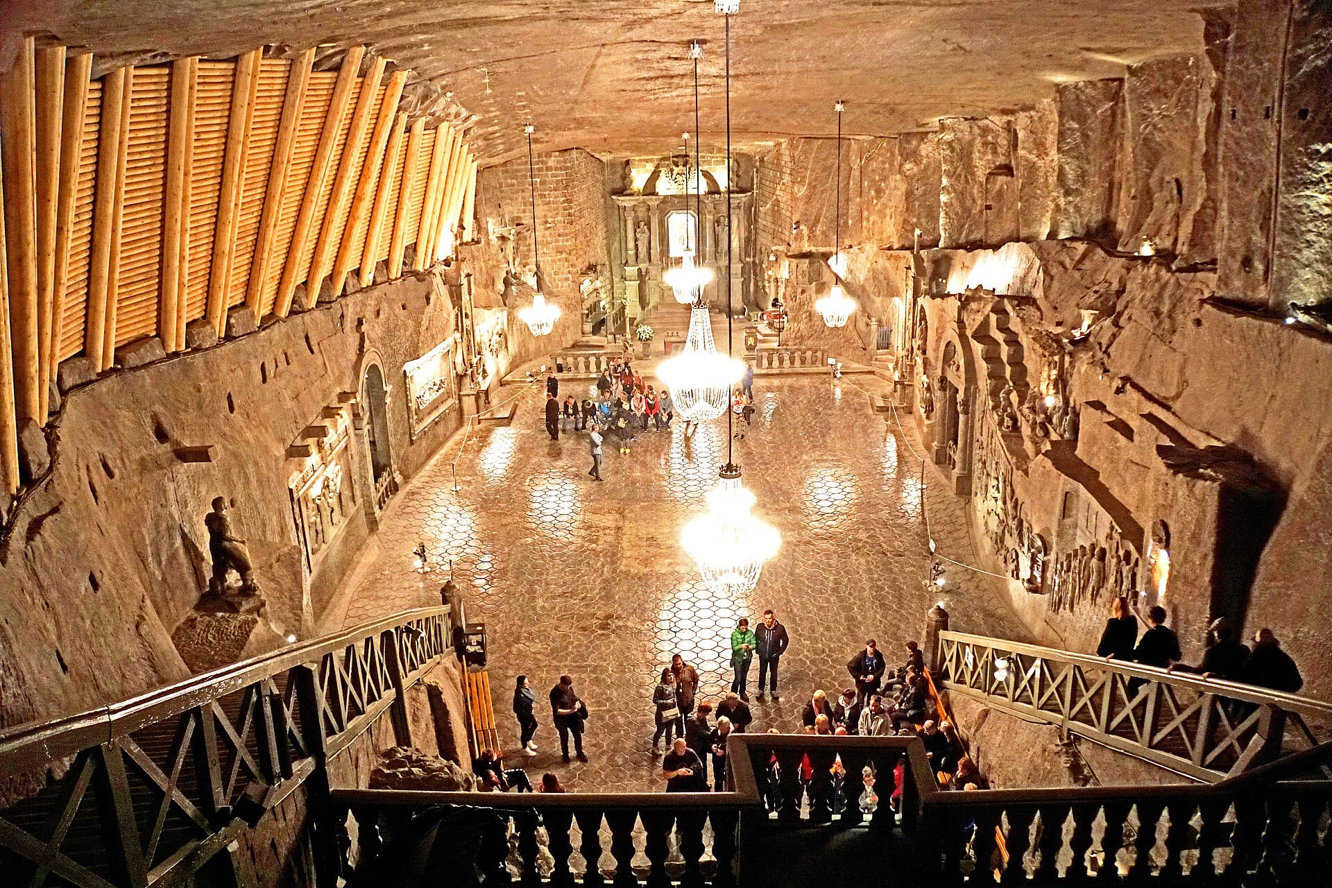 ห้องใต้ดินขนาดใหญ่ในเหมืองเกลือ Wieliczka พร้อมโคมระย้า ผนังเกลือแกะสลัก ประติมากรรม และกลุ่มนักท่องเที่ยวที่กำลังสำรวจพื้นที่