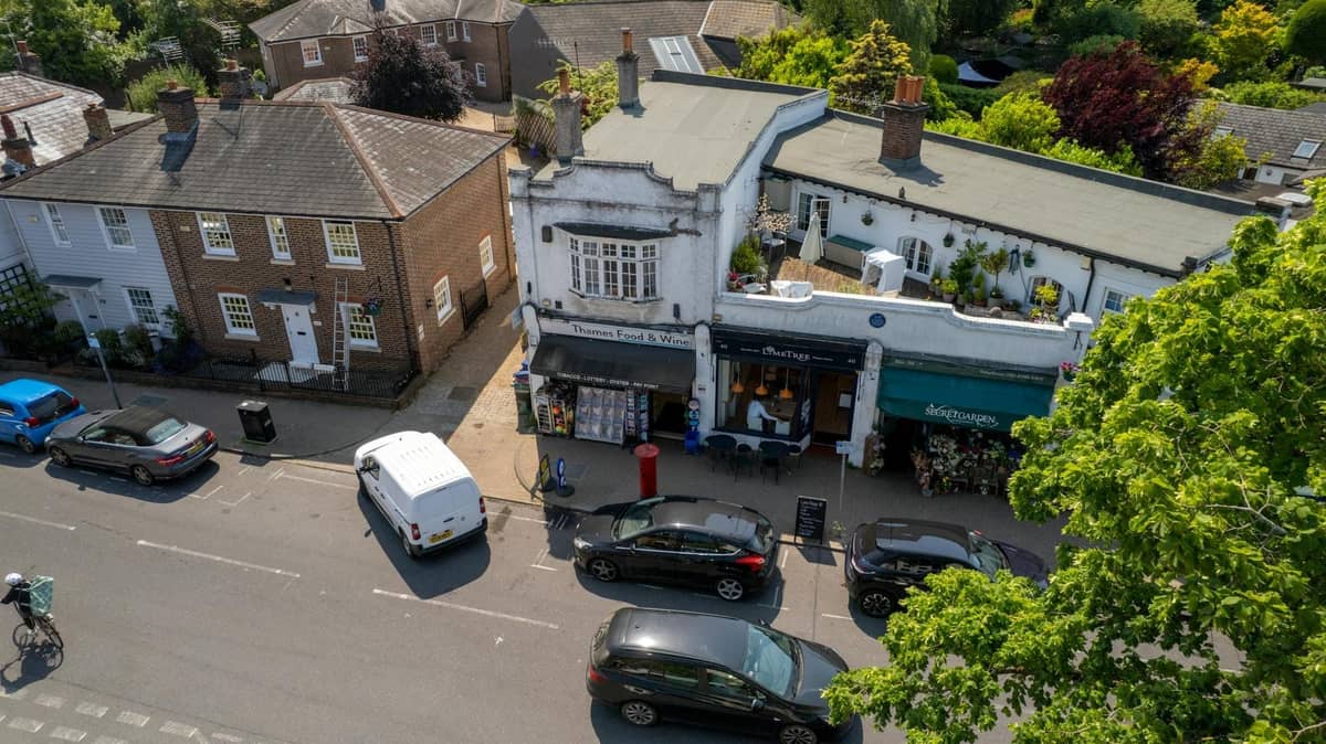 Vista aérea de uma agradável rua comercial de Londres com pequenas lojas independentes, uma banca de flores, carros estacionados e arredores arborizados, sugerindo uma atmosfera de aldeia.