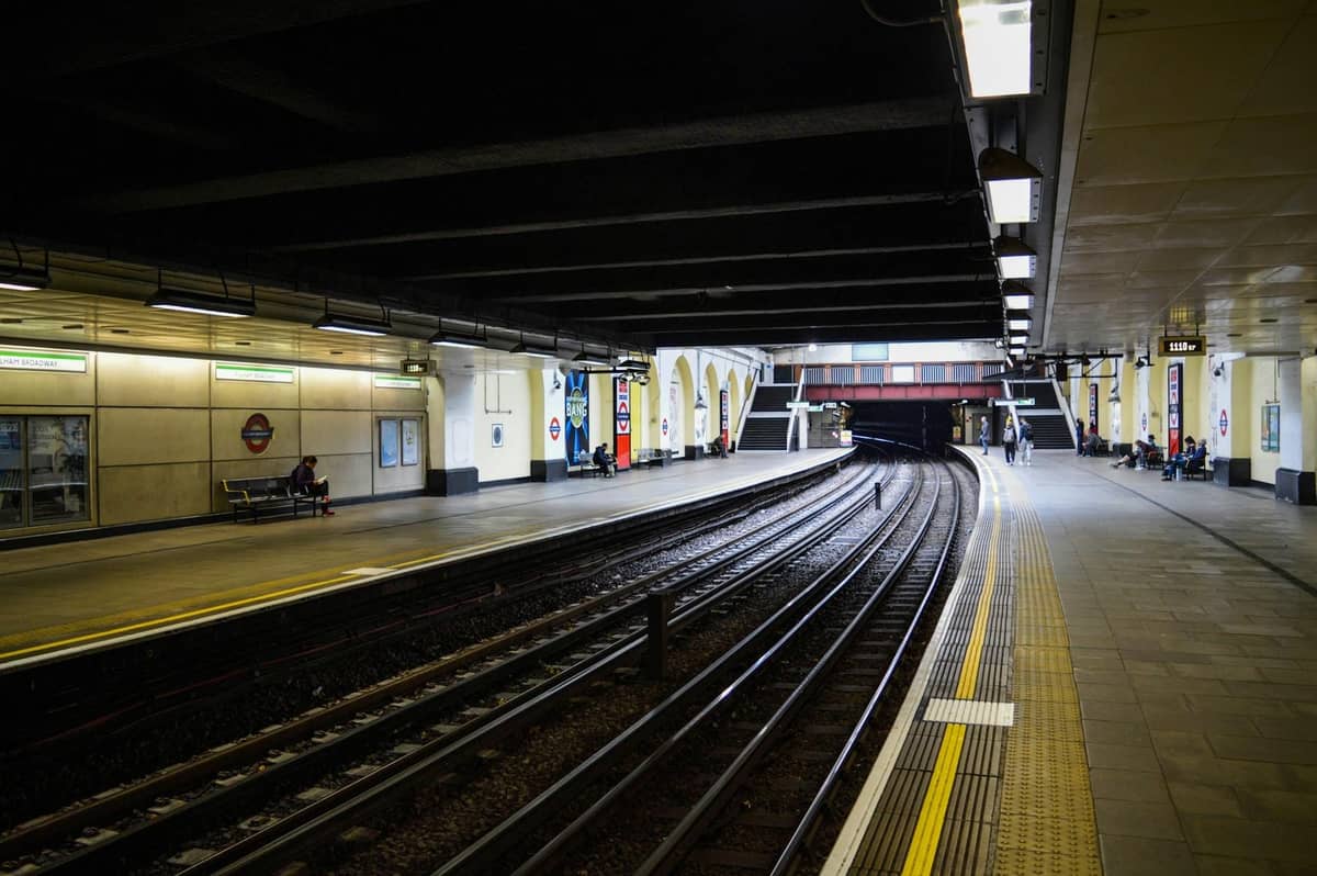 Plataforma da linha District do London Underground com trilhos visíveis e o característico roundel, mostrando uma cena real do transporte de Londres.