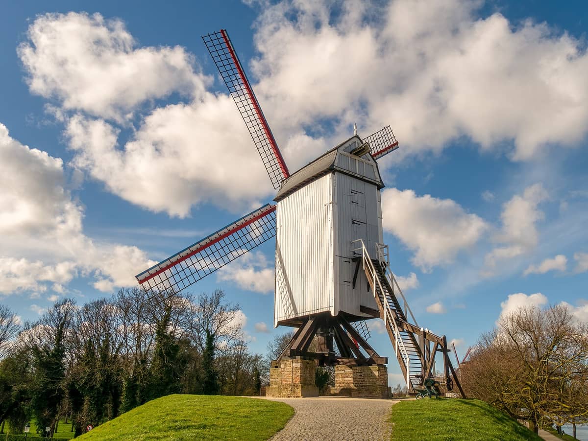 A traditional wooden windmill with red and white blades standing on grassy ramparts at Kruisvest in Bruges, under a blue sky with clouds.