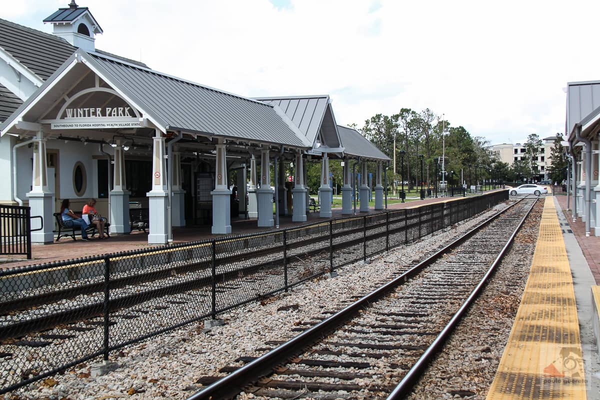 Winter Park train station platform with shelter, benches, and people waiting, surrounded by trees and tracks under a sunny sky.