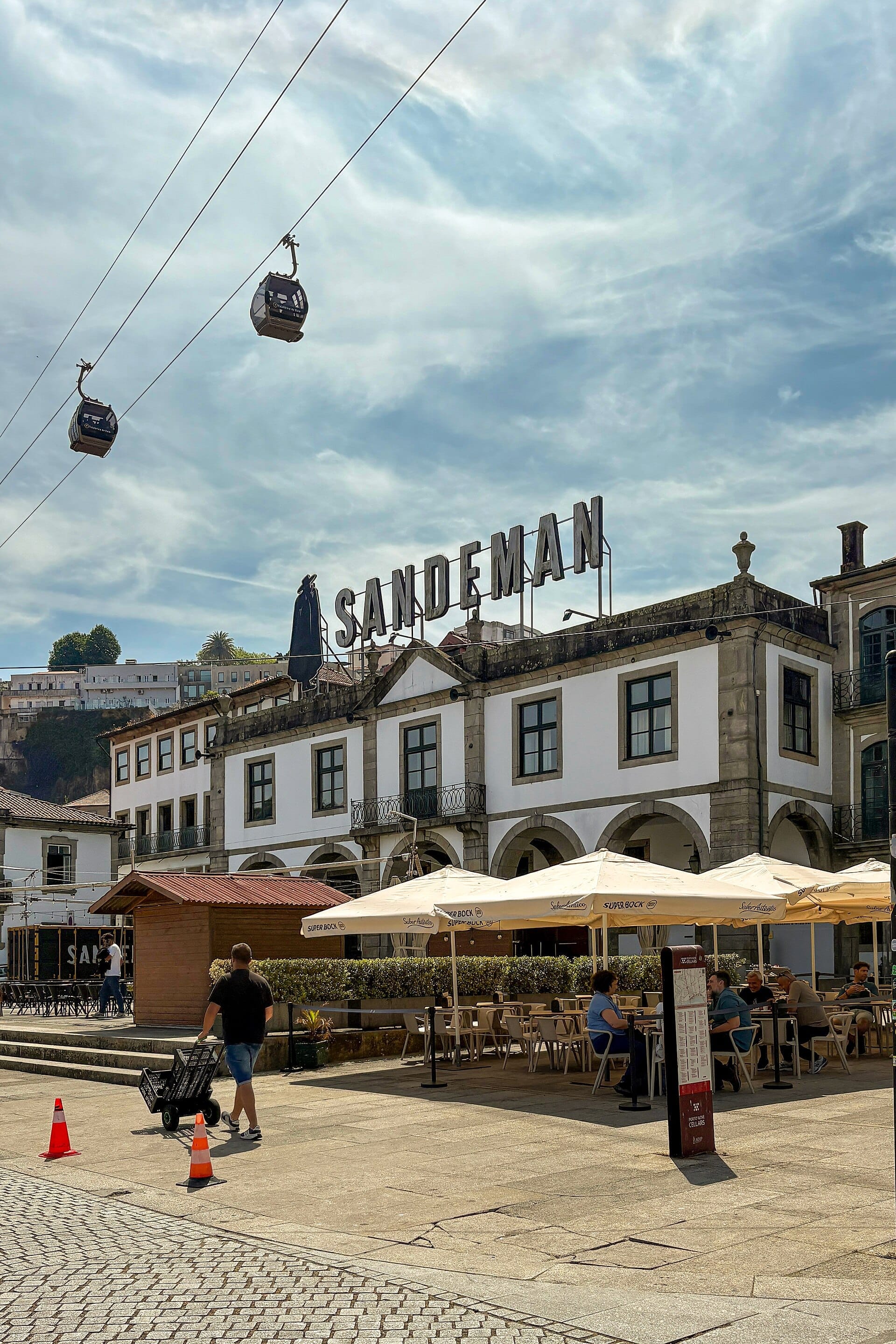Area pranzo all'aperto con commensali sotto ombrelloni bianchi davanti all'edificio Sandeman al WOW Porto, con funivie che passano in alto.