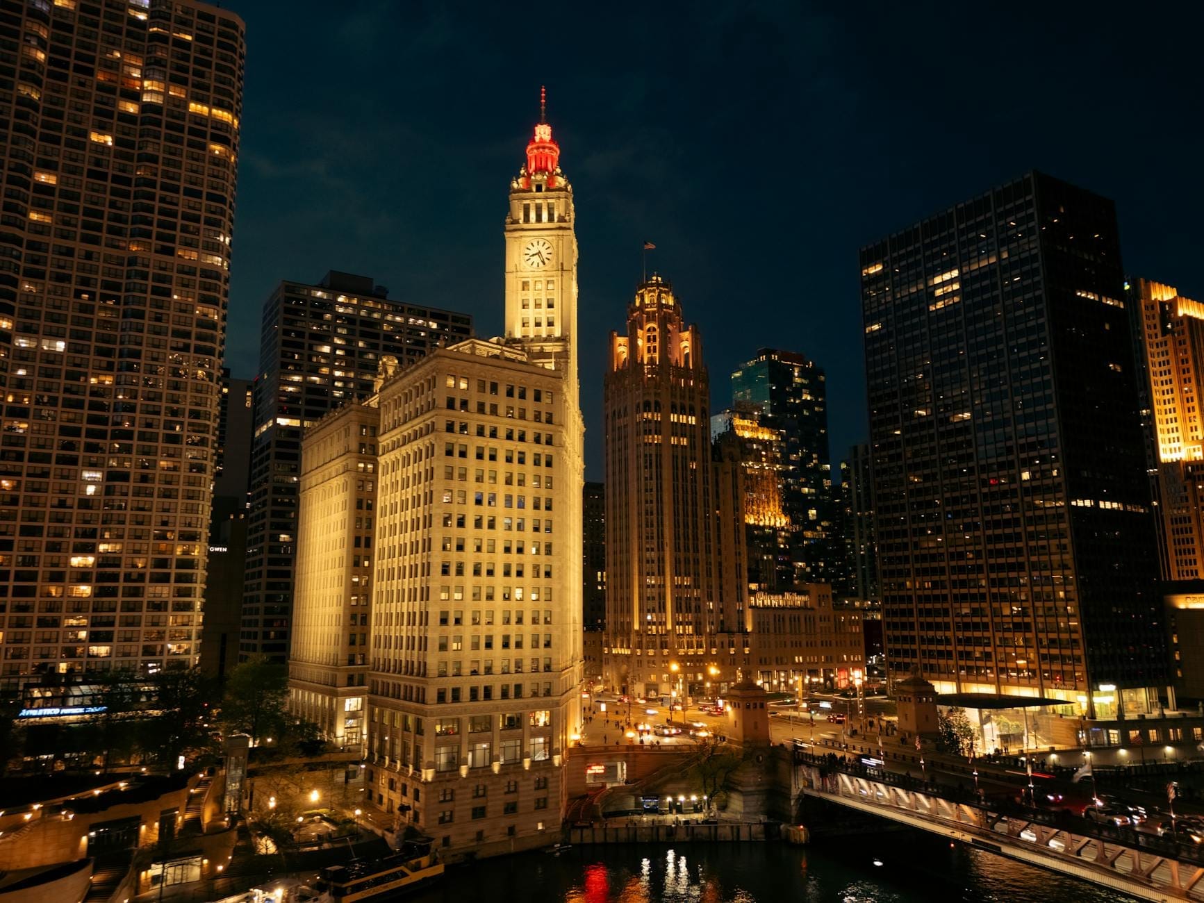 Vista notturna del Wrigley Building illuminato con la sua torre dell'orologio, circondato dai grattacieli di Chicago e dal Chicago River in basso, in un vivace panorama urbano.