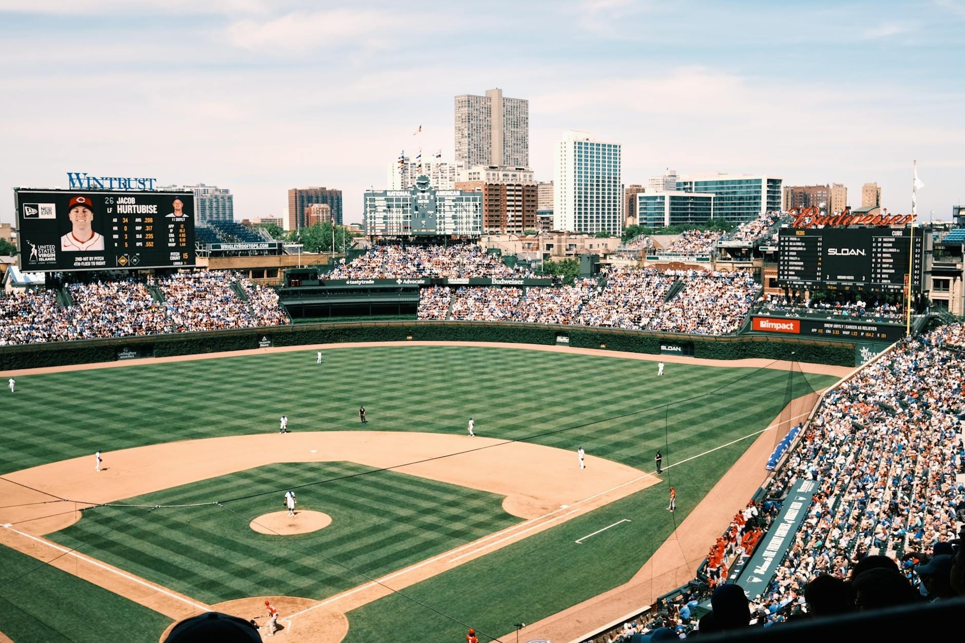Vista panoramica di Wrigley Field durante una partita diurna, con il diamante verde, le tribune gremite e lo skyline di Chicago sullo sfondo.