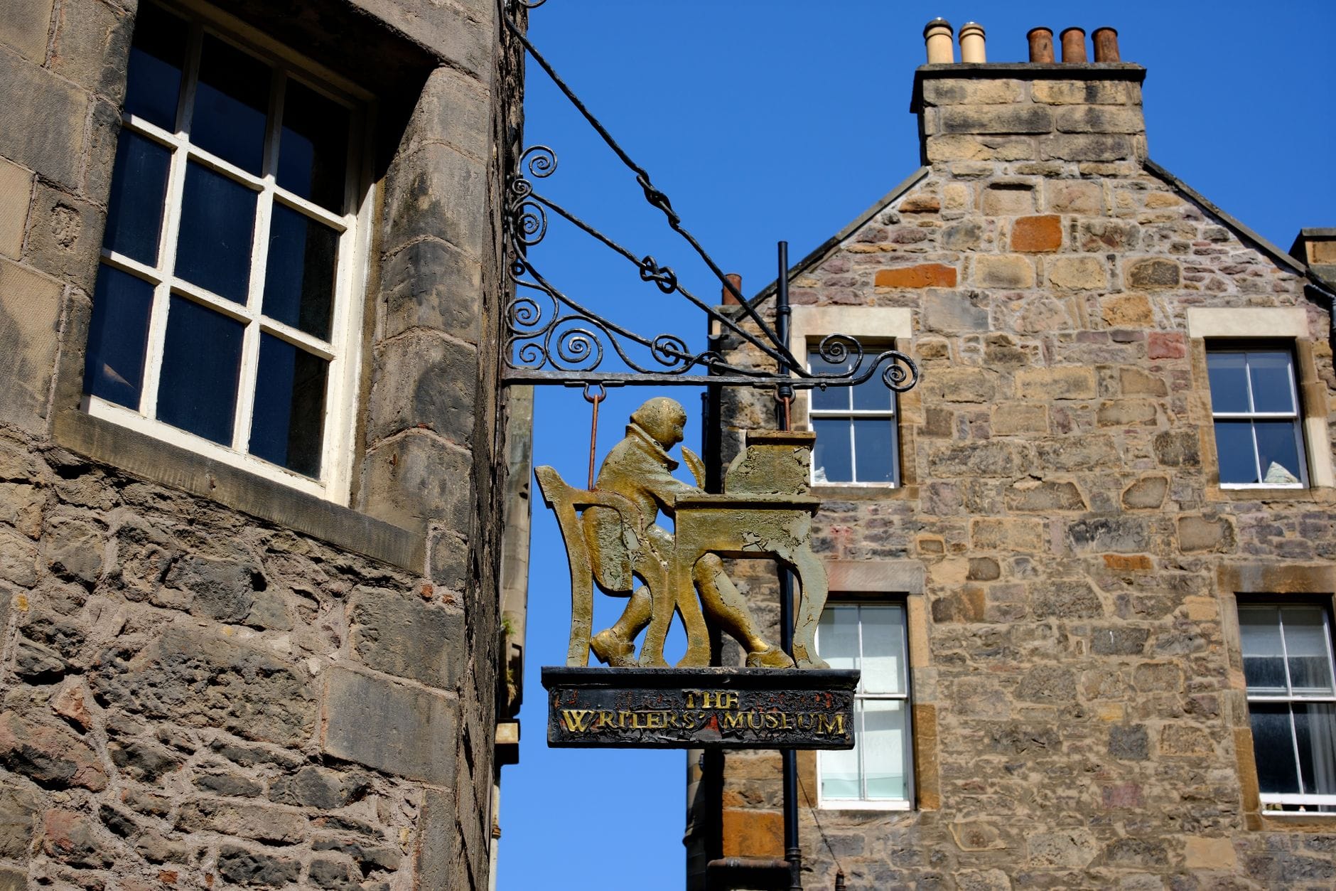 Verziertes Hängeschild des Writers' Museum in Edinburgh mit einer goldenen Figur an einem Schreibtisch, angebracht an einem historischen Steingebäude an einem sonnigen Tag.