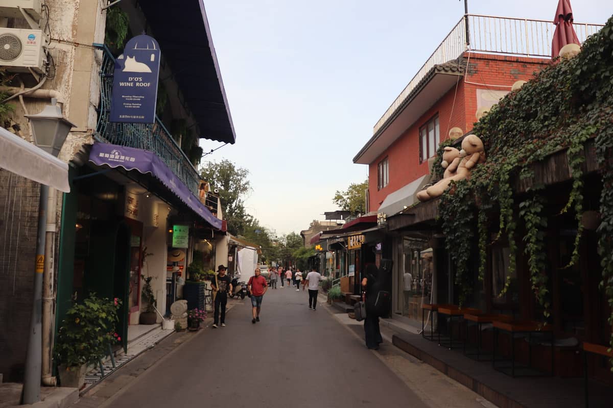 Ein entspannter Blick in den Wudaoying Hutong in Peking: Menschen schlendern vorbei, kleine unabhängige Läden reihen sich aneinander, begrünte Fassaden und Tische im Freien säumen die Gasse.